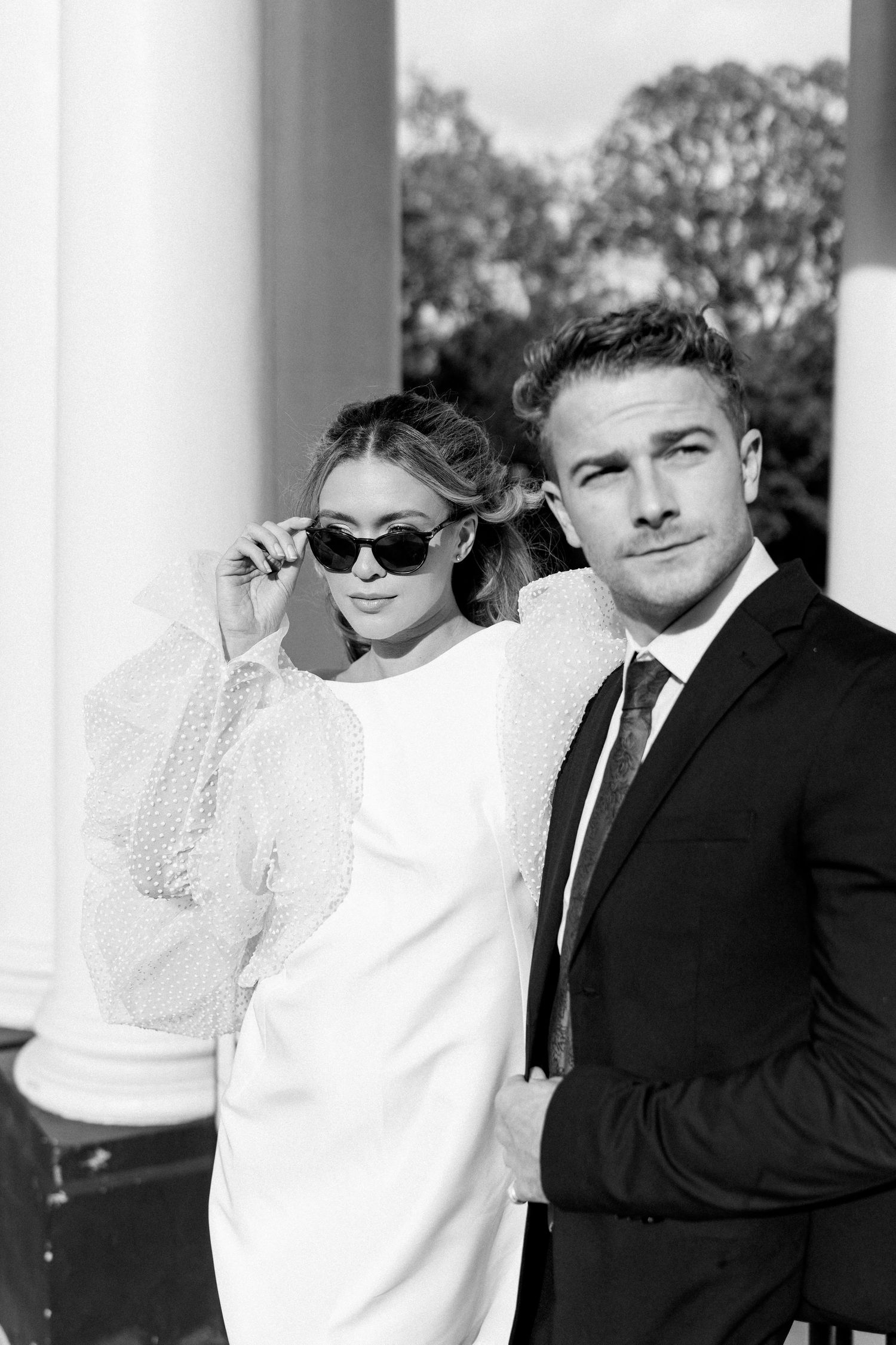 Stylish couple poses against white pillars in black and white photography.