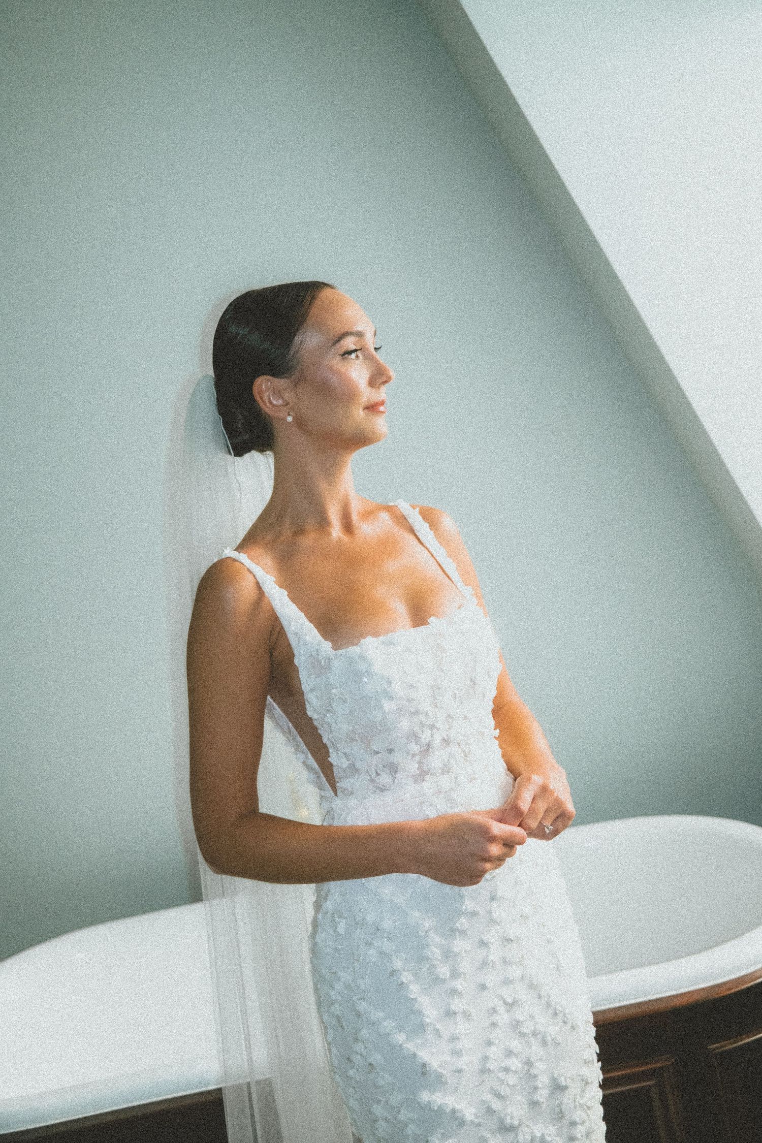 A bride in a fitted white lace gown and veil stands in profile against a light wall in soft natural lighting.