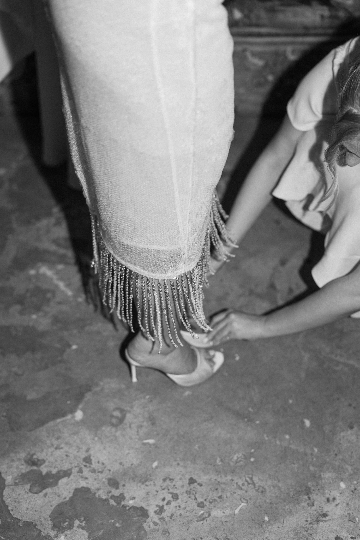 Black and white photo of bride in fringed wedding dress having her heels adjusted.