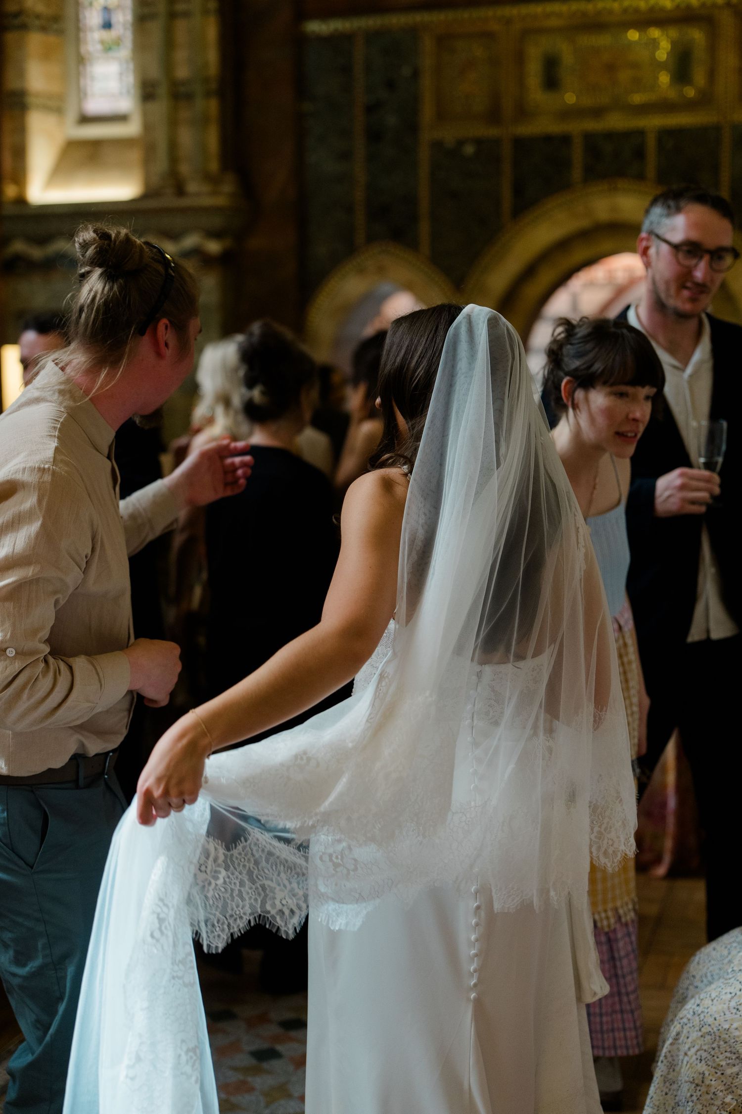 Wedding assistant helps bride with lace gown and veil inside ornate church surrounded by guests.