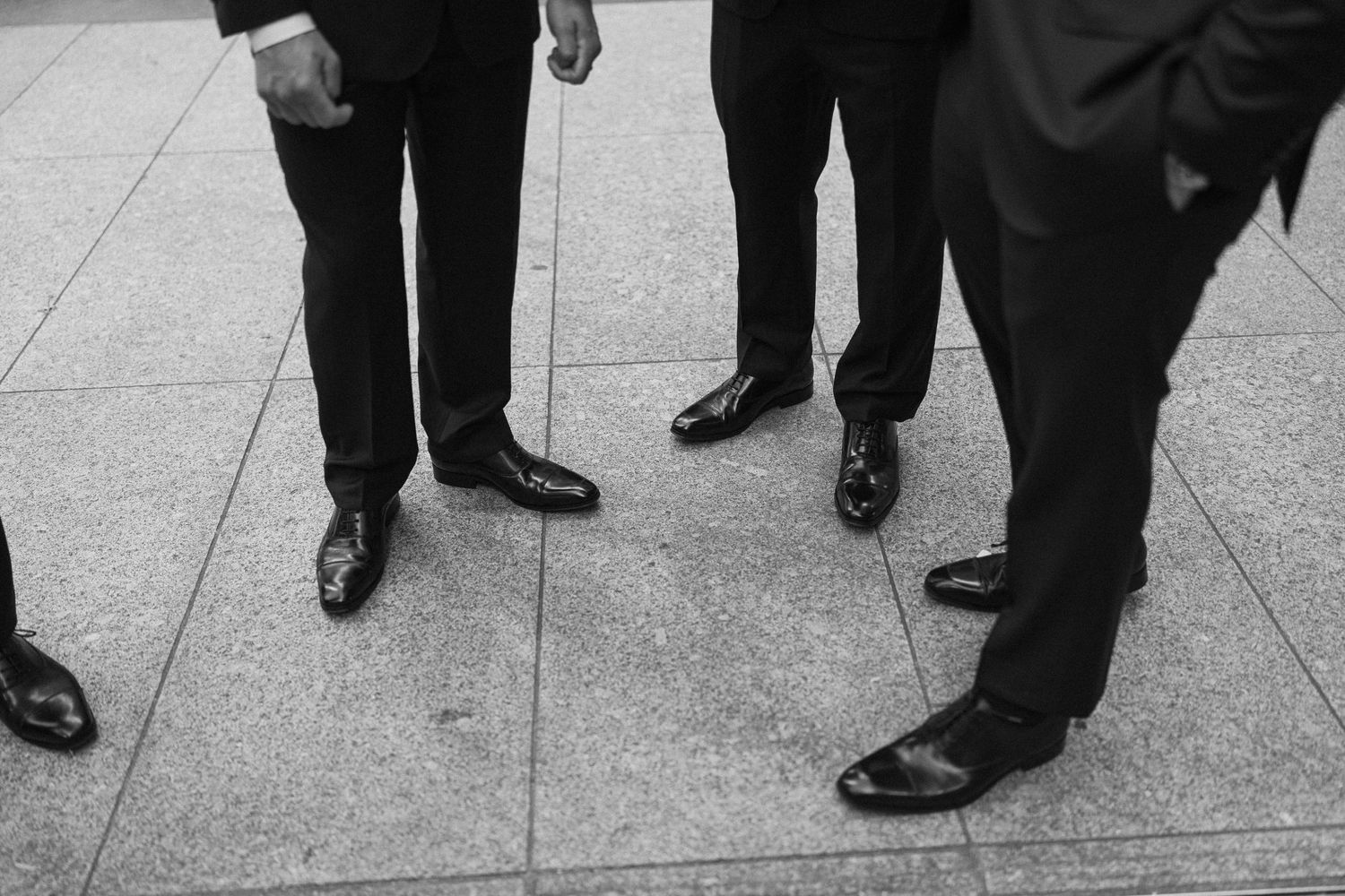 Black and white photo of groomsmen in formal suits and dress shoes standing on tiled pavement at a wedding.