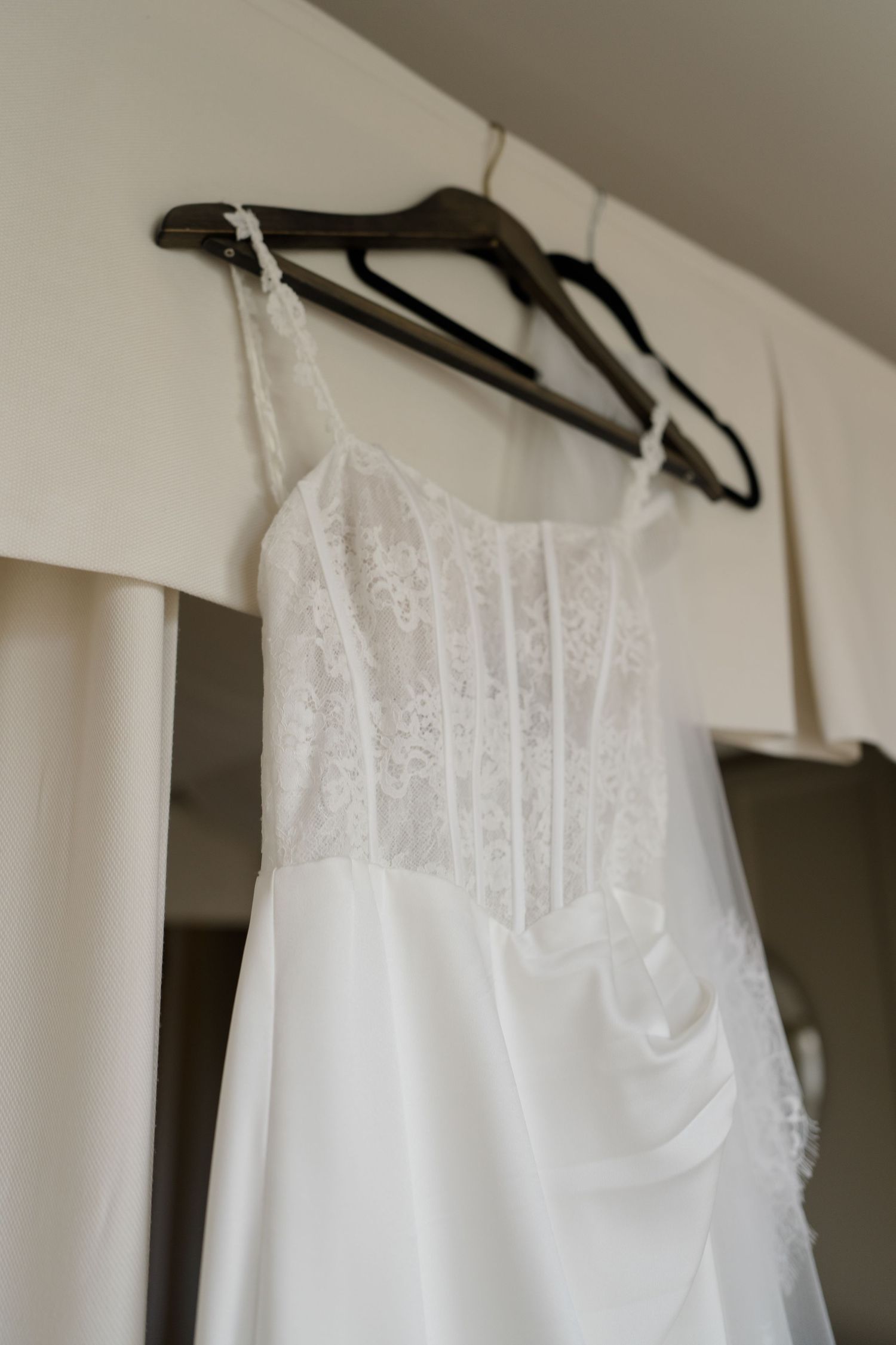 Elegant white lace and satin wedding dress hanging on dark hangers against a light wardrobe in a bridal suite.