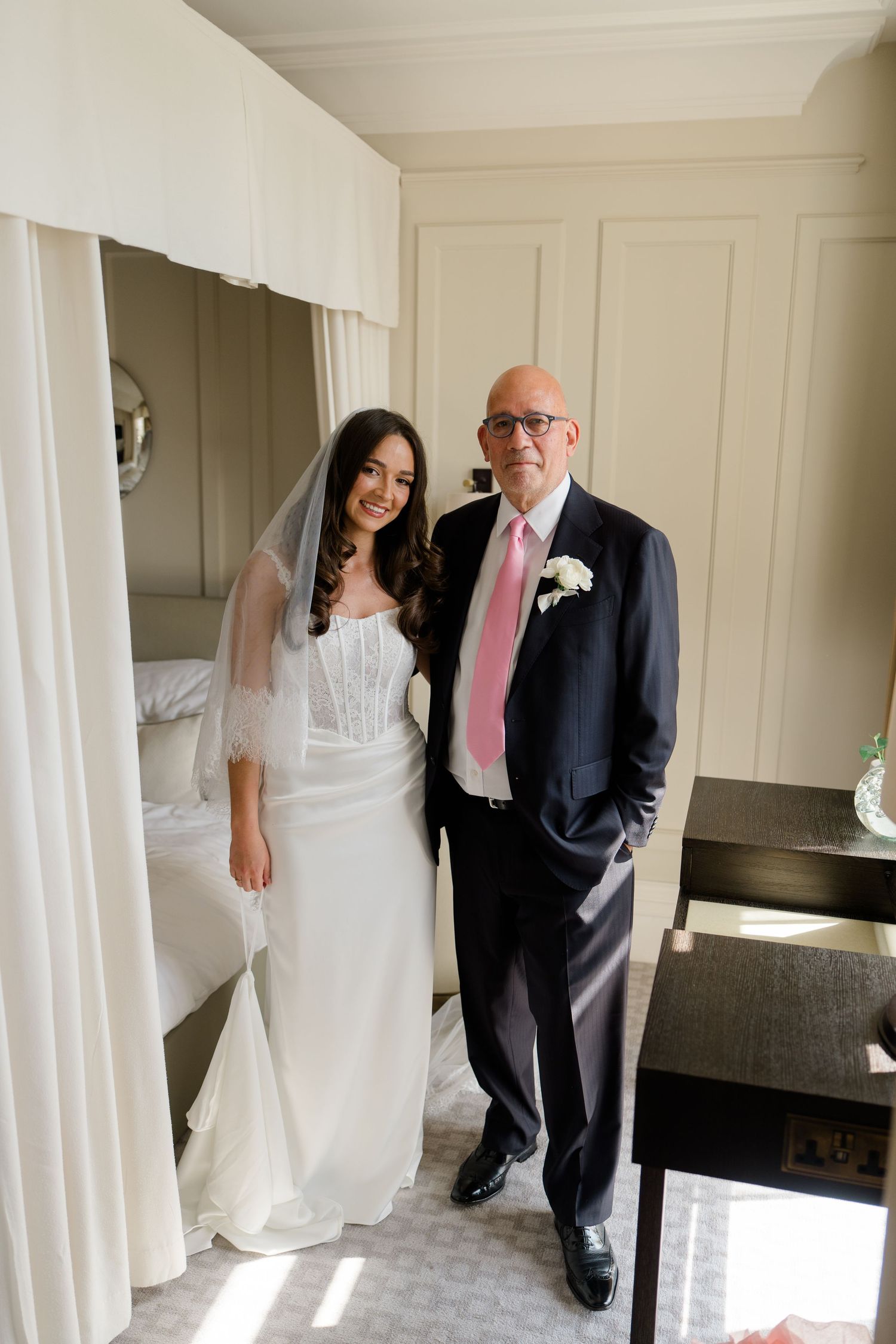 Bride in white wedding dress and veil posing with her father in a suit and pink tie.