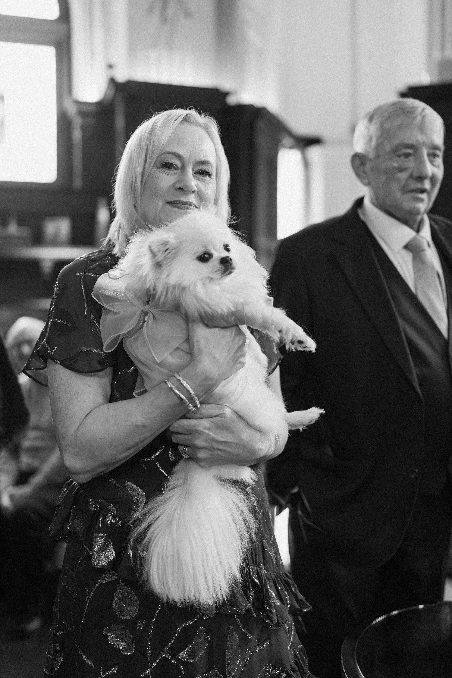 Smiling blonde woman holding a fluffy white Pomeranian dog at a formal indoor event.