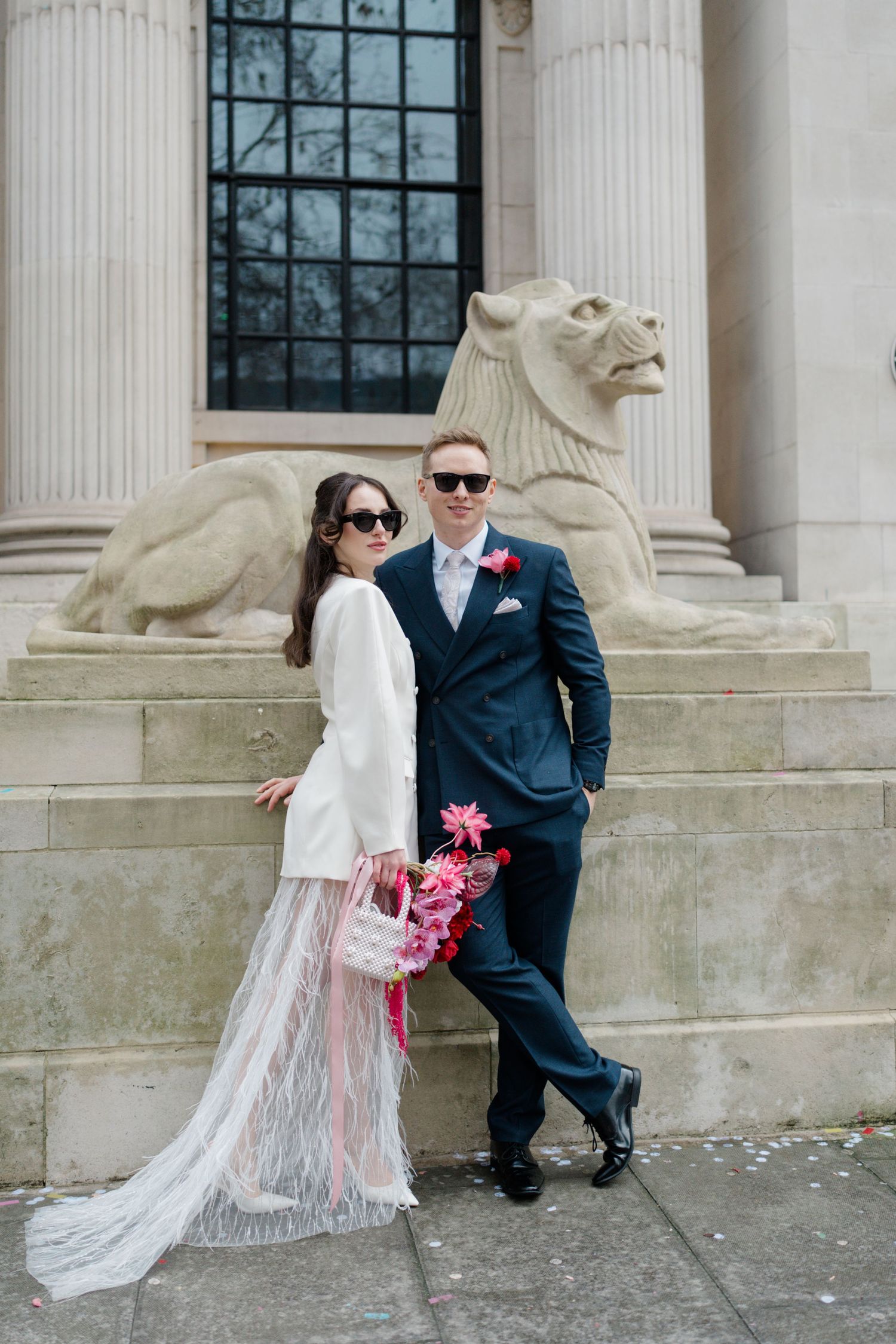 Bride in white blazer and feather skirt poses with groom before stone lion sculpture outdoors.