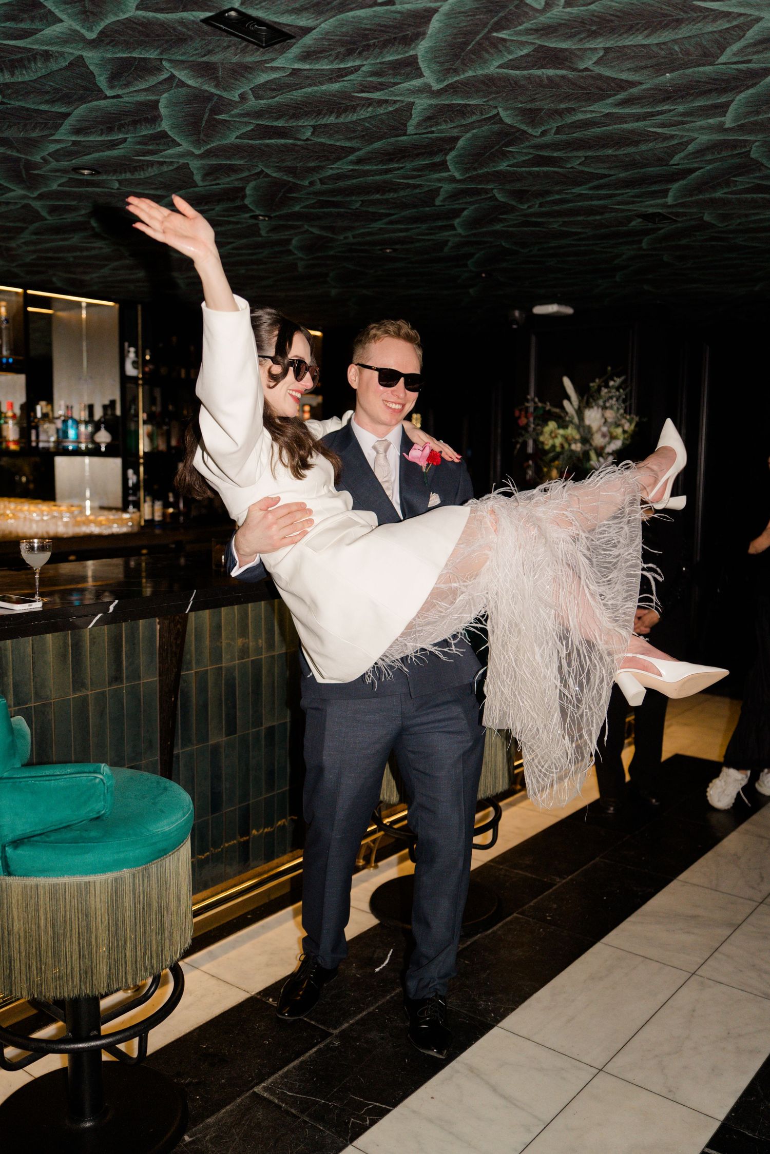 Groom carrying laughing bride in white feather wedding dress at stylish bar venue with green decor.