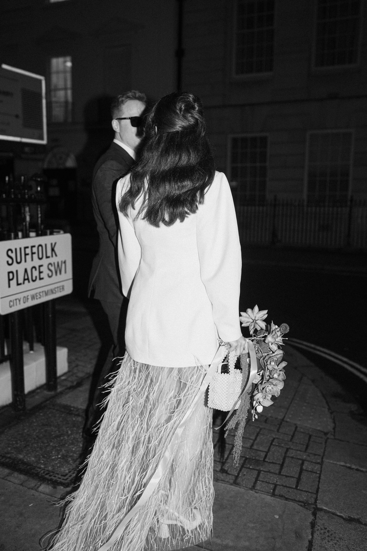 Black and white night photo of couple near Suffolk Place SW1 sign, bride in white feather skirt.