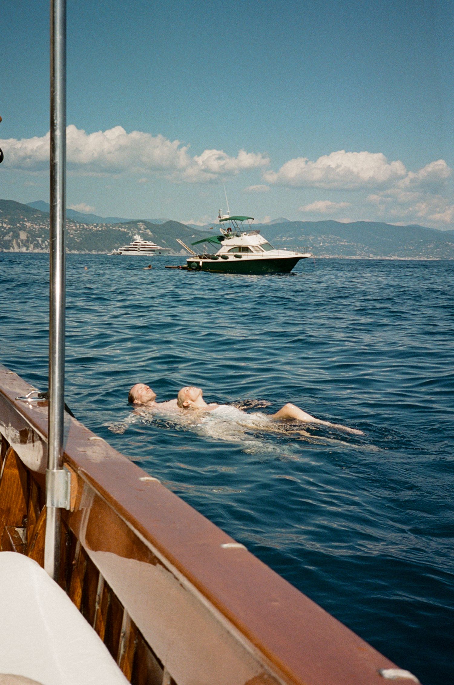 Two swimmers enjoy the blue Mediterranean sea near a wooden yacht with a motor boat and mountains in the background.