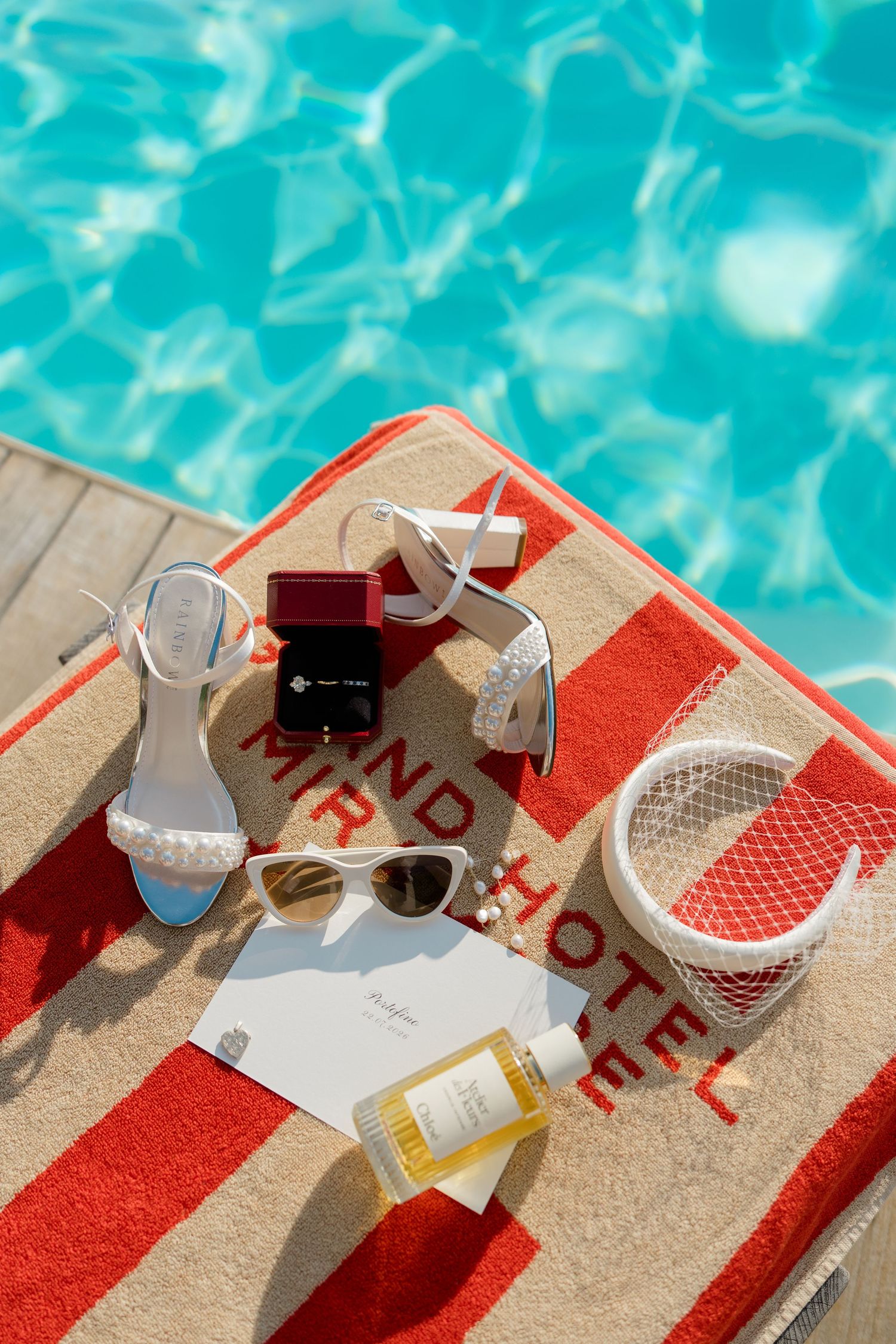 White heeled sandals, sunglasses, ring box, and perfume on a striped Grand Hotel towel by a turquoise pool.