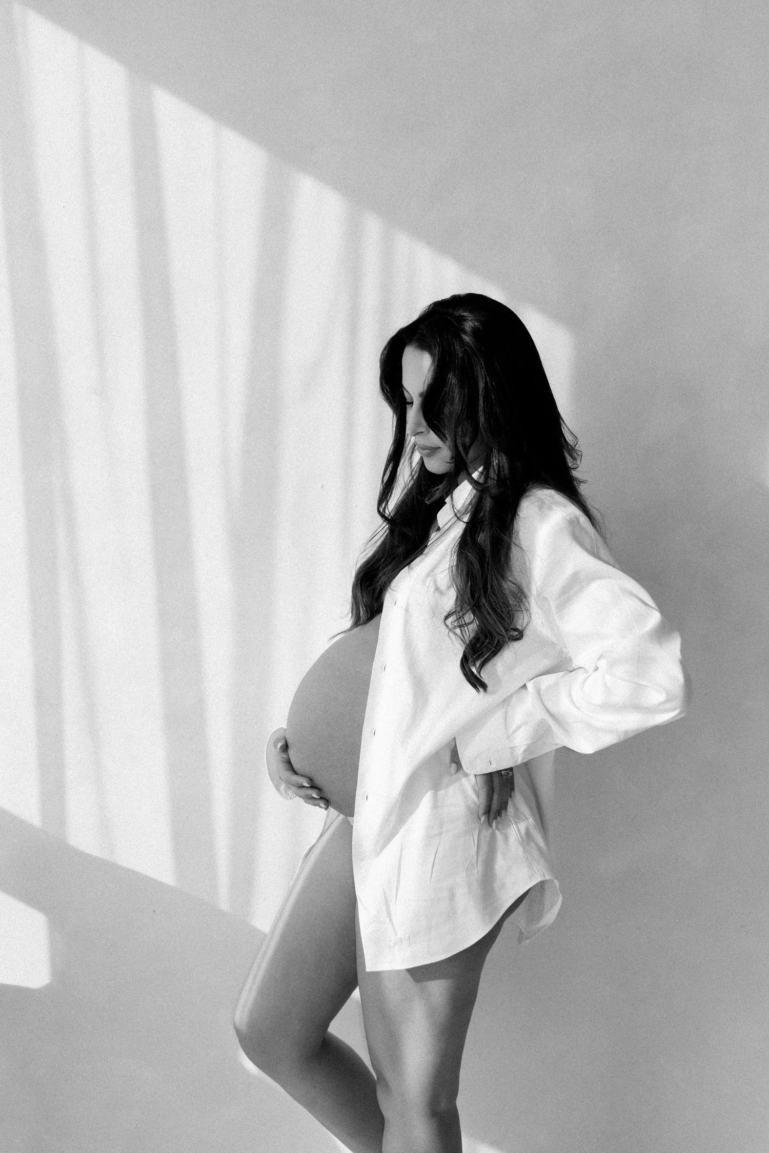 Black and white photo of a pregnant woman in an open white shirt cradling her baby bump in soft natural light.