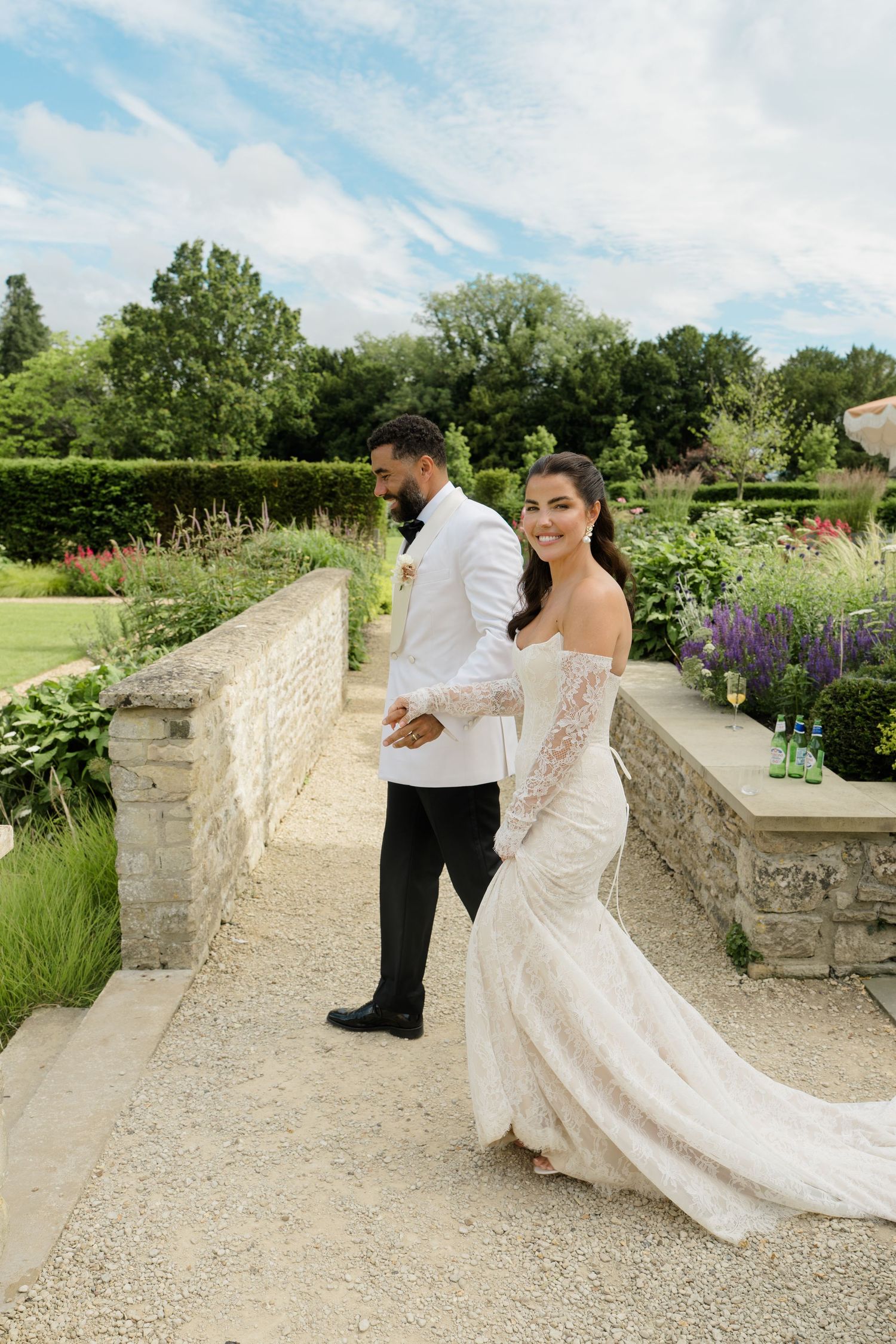 Bride in lace gown holds grooms hand walking through manicured garden at English countryside wedding.