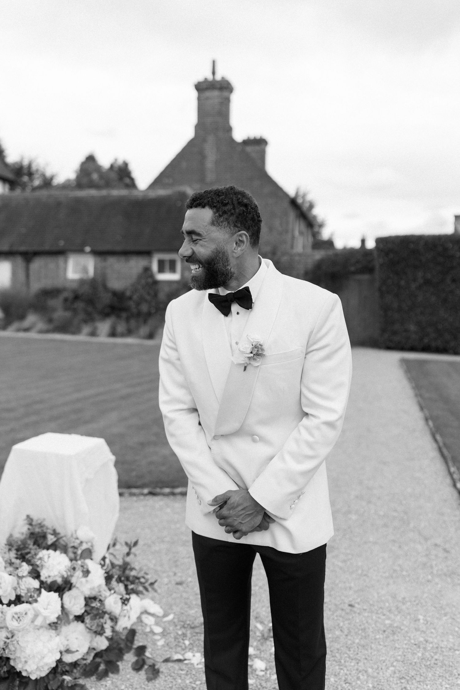 Groom in white tuxedo jacket and black bow tie smiling outdoors at elegant wedding venue.
