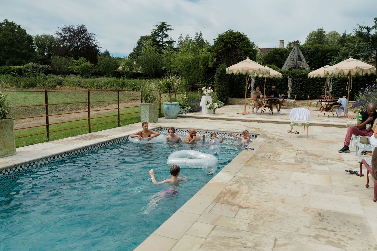 Wedding guests enjoying a sunny outdoor pool party with inflatables and straw umbrellas at a manor.