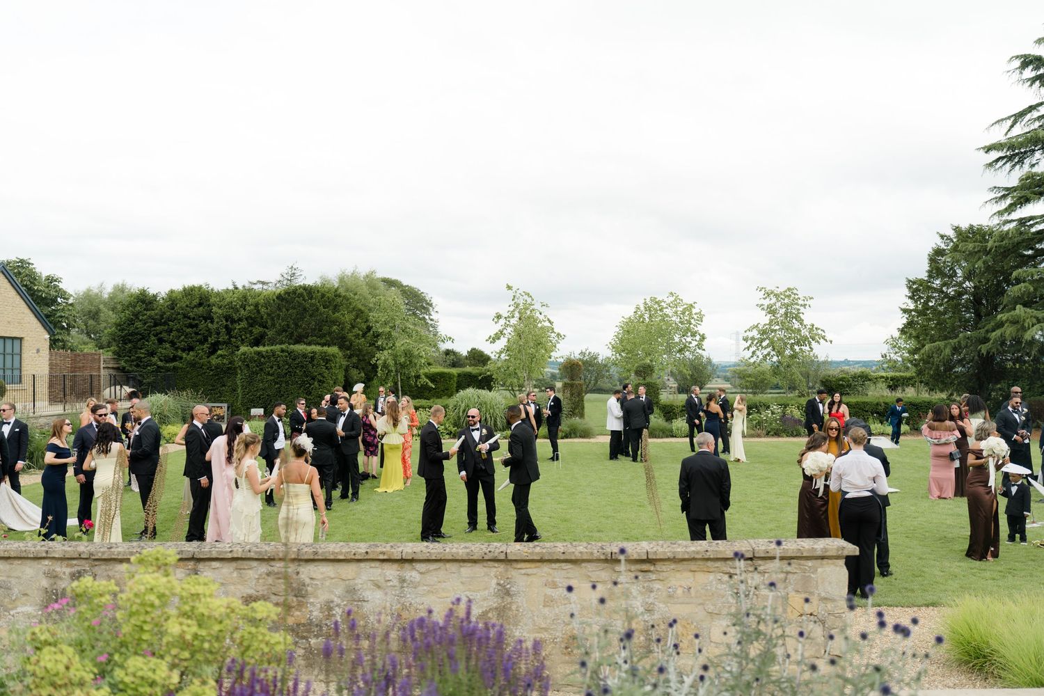 Wedding guests mingling on a manicured garden terrace with hedgerows and rolling countryside views.