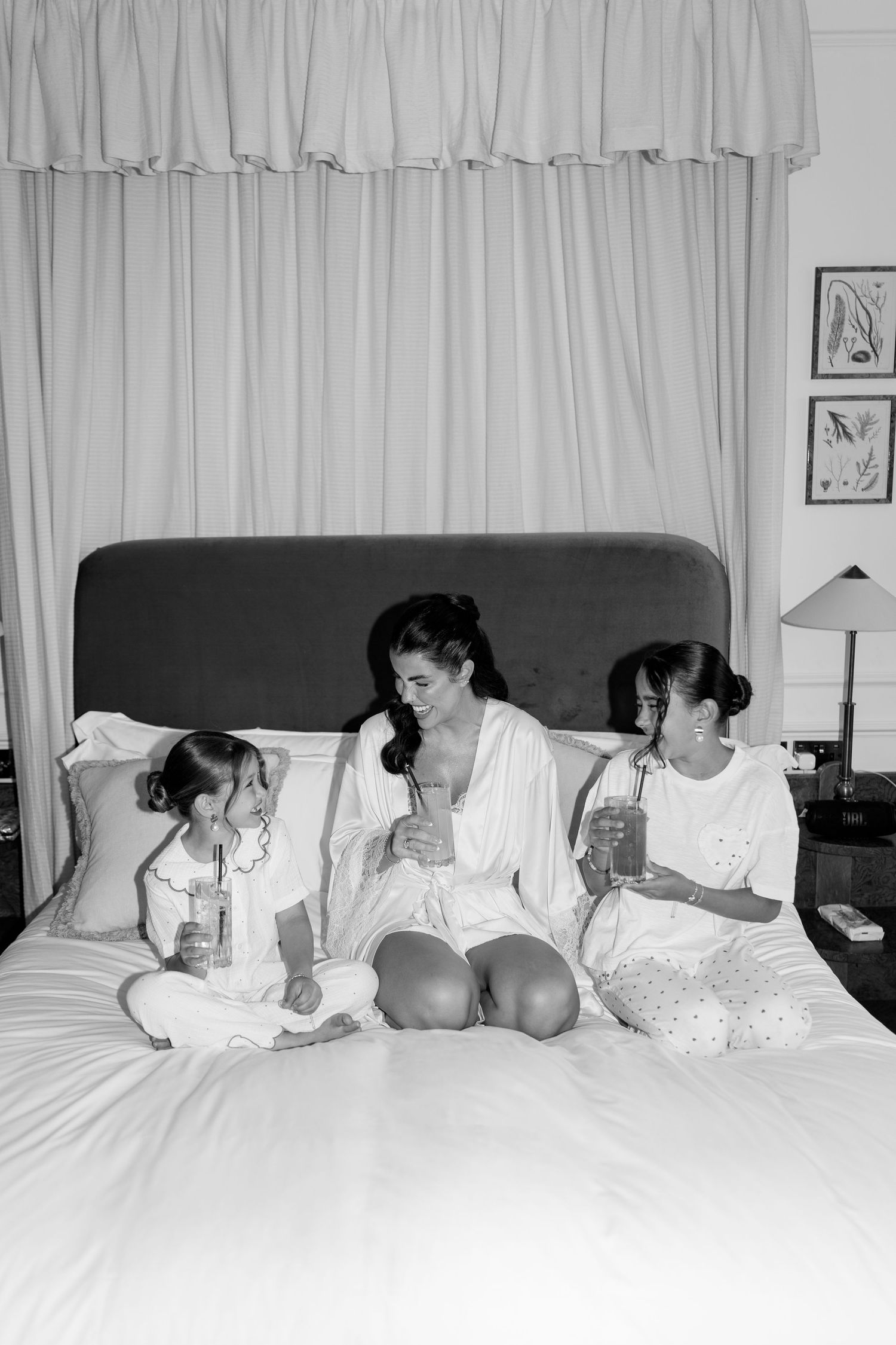 Black and white photo of bride in robe sitting on bed with two young girls drinking smoothies.