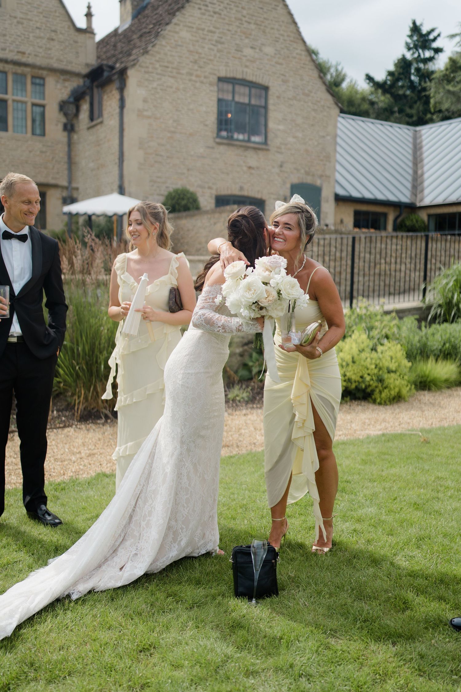 Bride in lace gown embraces guest while bridesmaids in yellow dresses stand nearby on lawn.