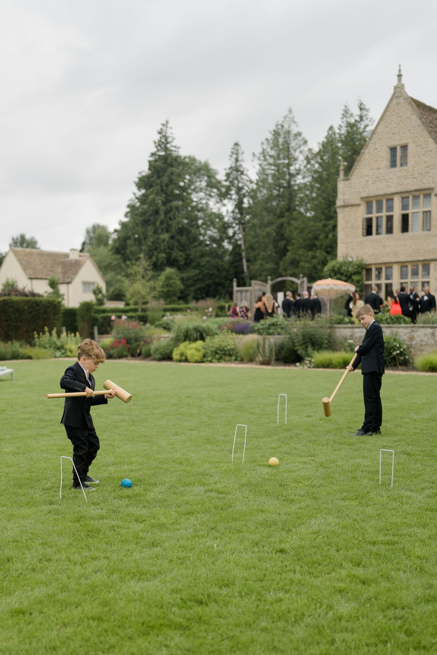 Two boys in black suits play croquet on a manicured lawn at a countryside wedding venue.