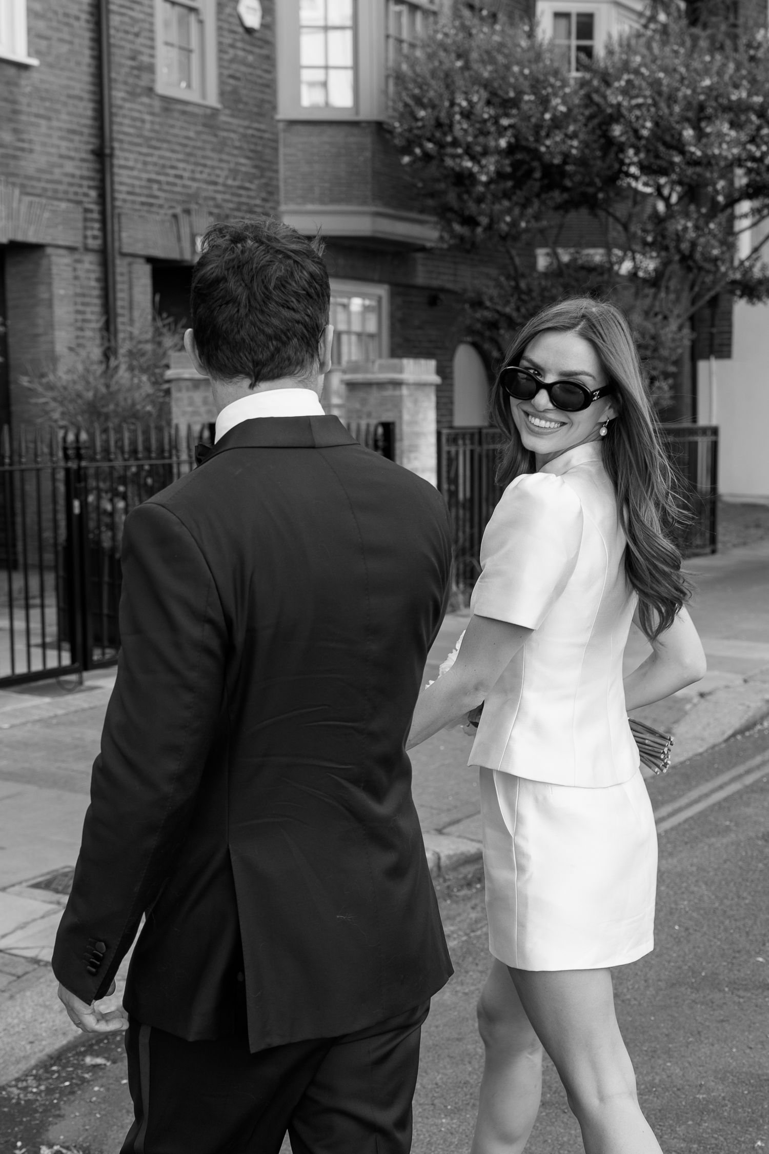 Black and white photo of an elegant couple walking on a city street, woman in white mini dress and sunglasses glancing back.
