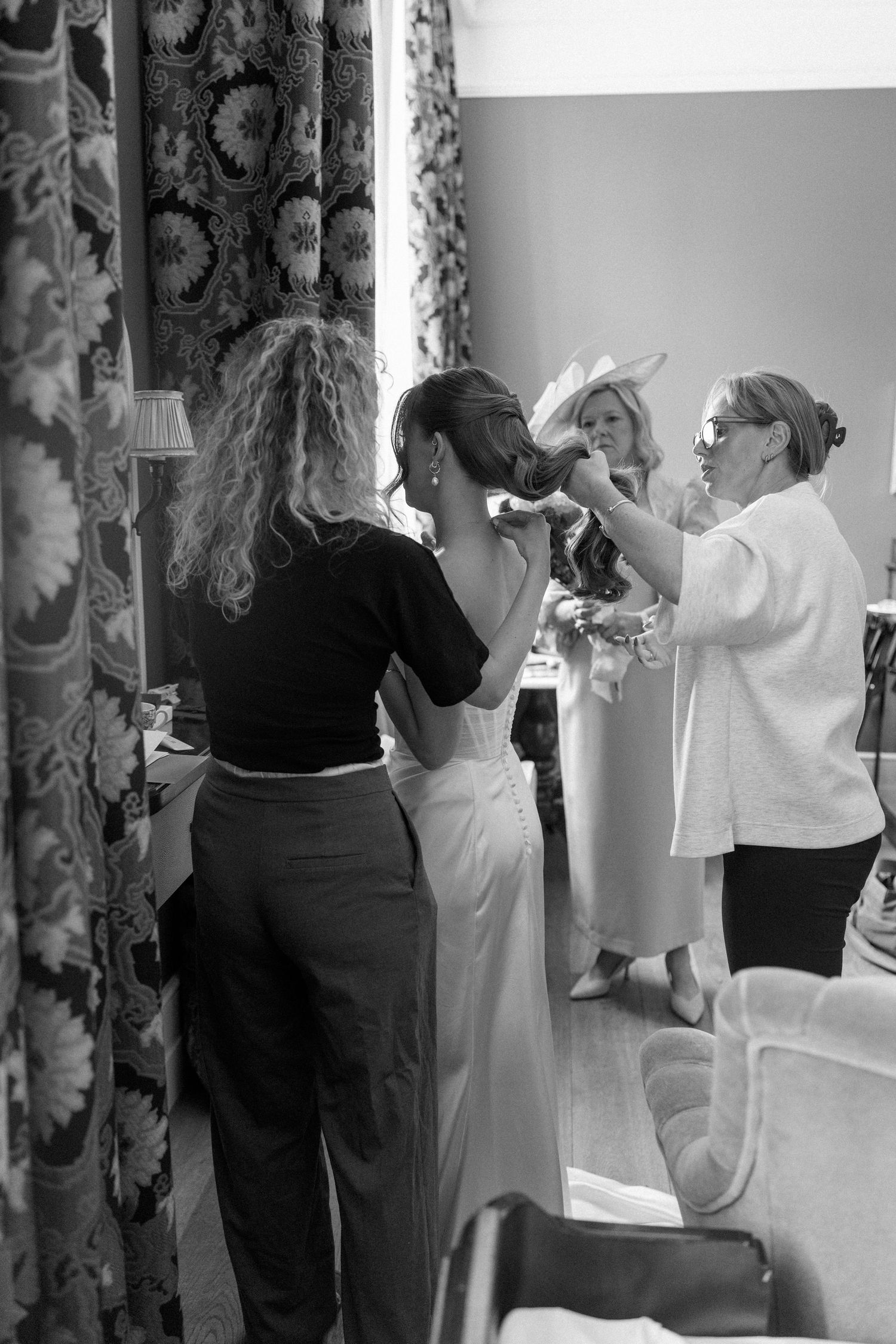 Black and white photo of a bride getting her hair styled by two women in an elegant room with floral curtains.