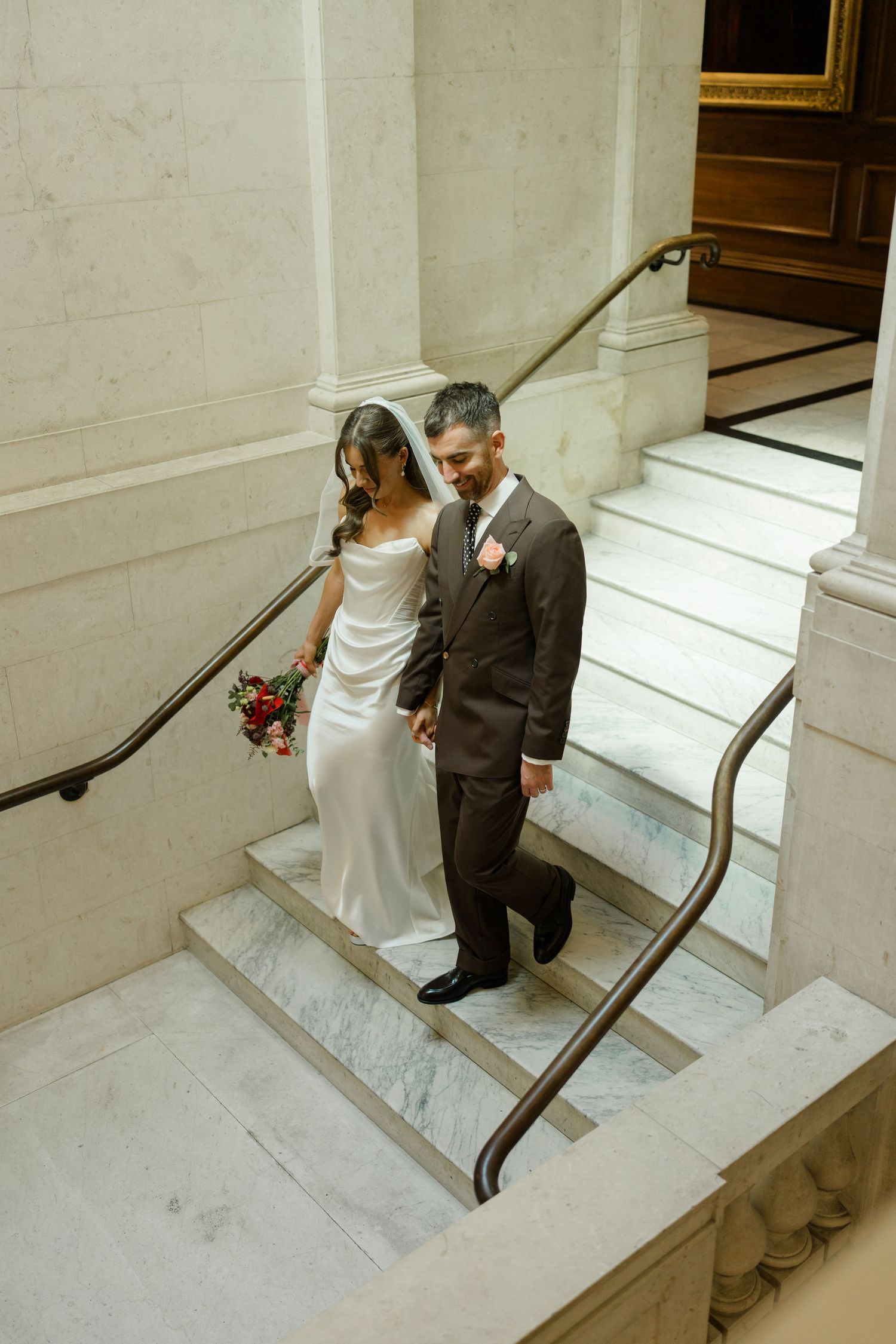 Bride and groom descending grand marble staircase together in elegant wedding venue.