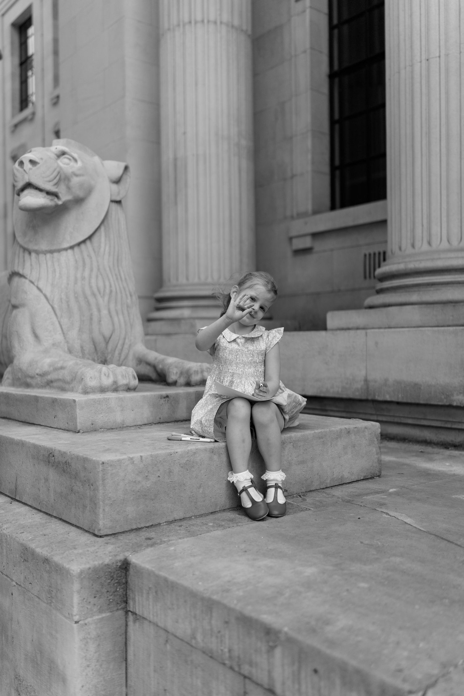 Black and white photo of young girl in dress sitting on steps beside stone lion sculpture.