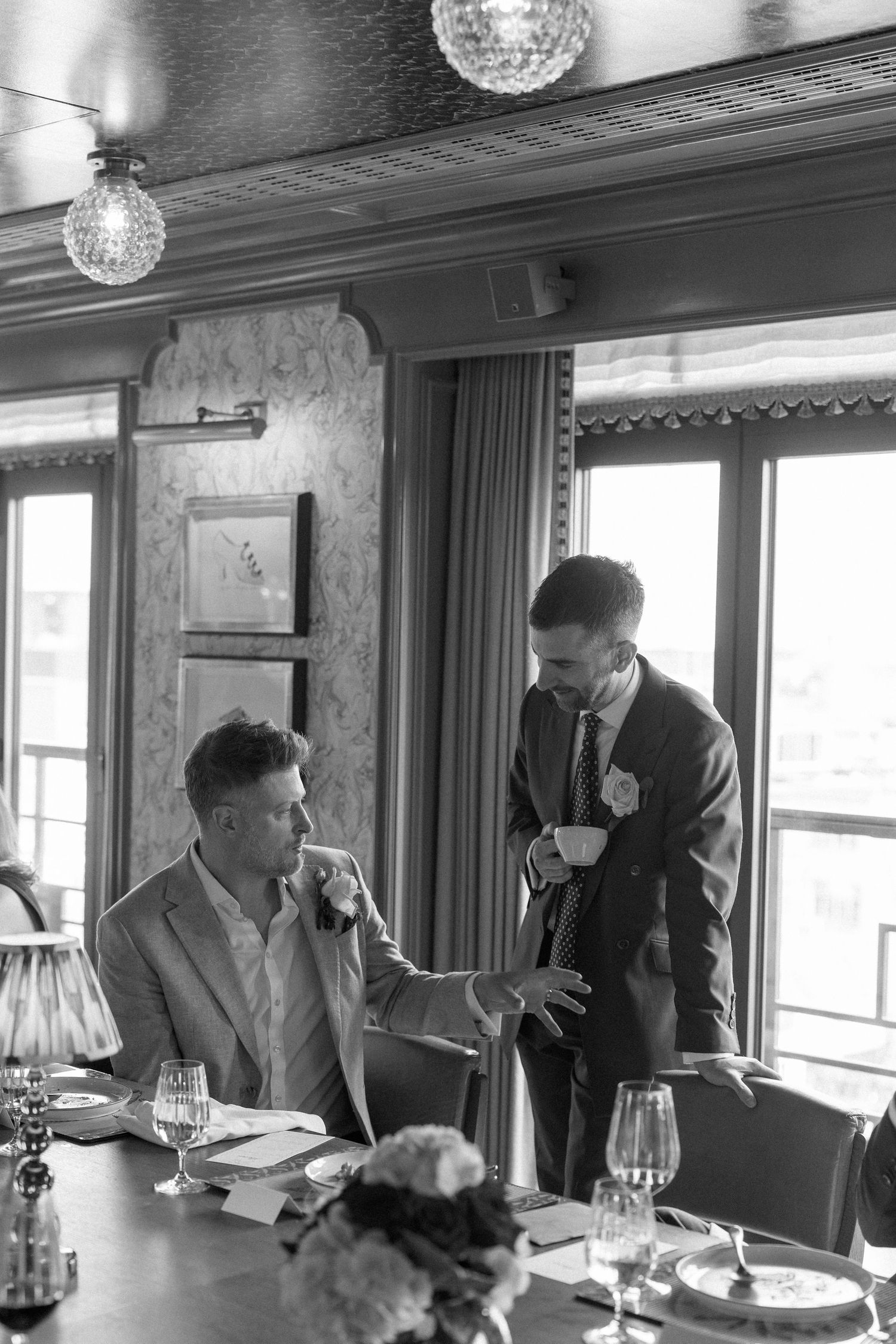Groom in dark suit with boutonniere speaks to seated guest at elegantly set wedding reception table.