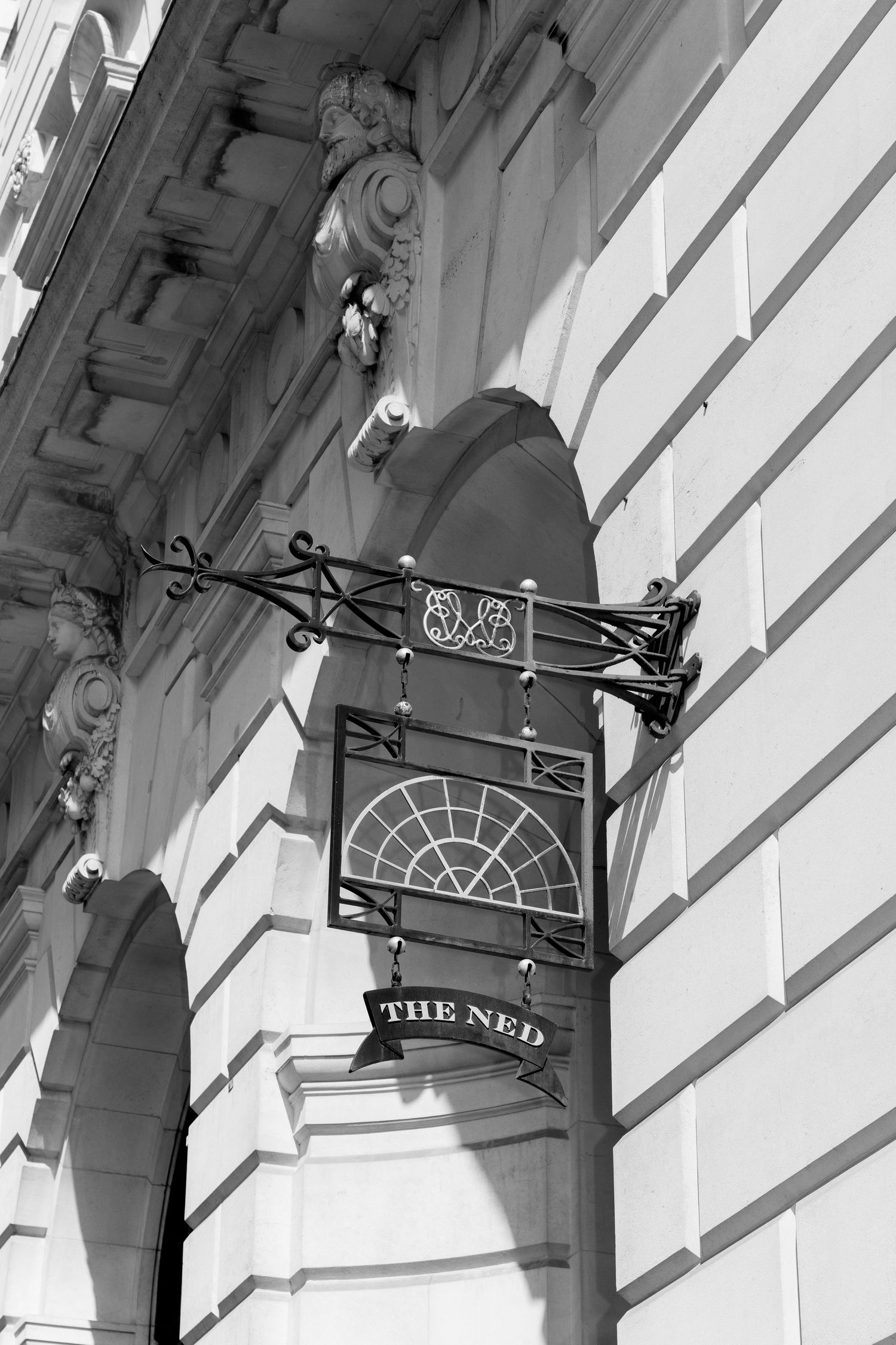 Black and white photo of The Ned hotel sign mounted on ornate stone building facade in London.