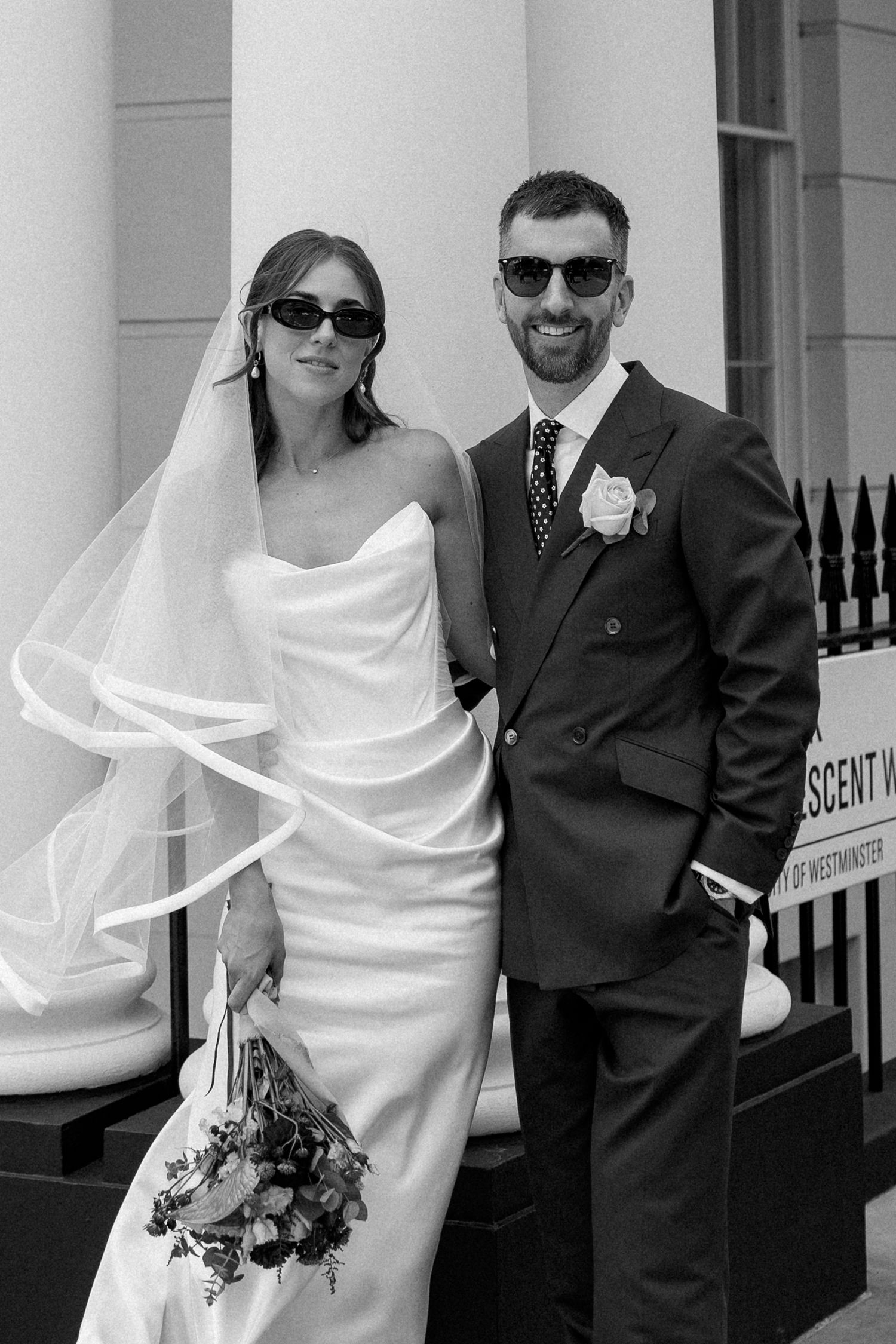 Stylish bride and groom wearing sunglasses pose together outside a white columned building on their wedding day.