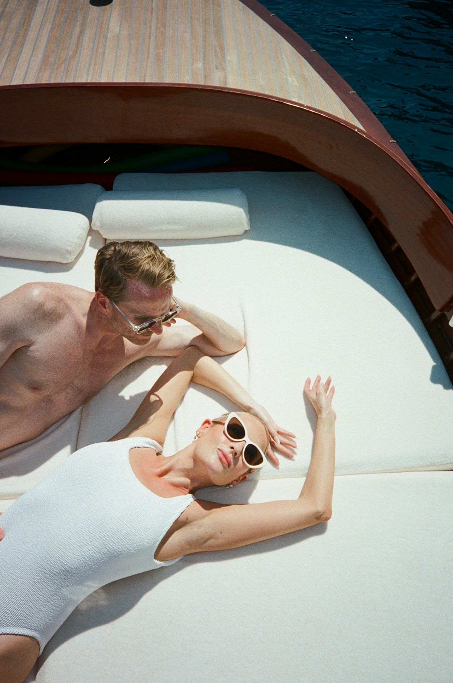 Couple sunbathing on a luxury wooden boat deck, both wearing sunglasses, viewed from above.