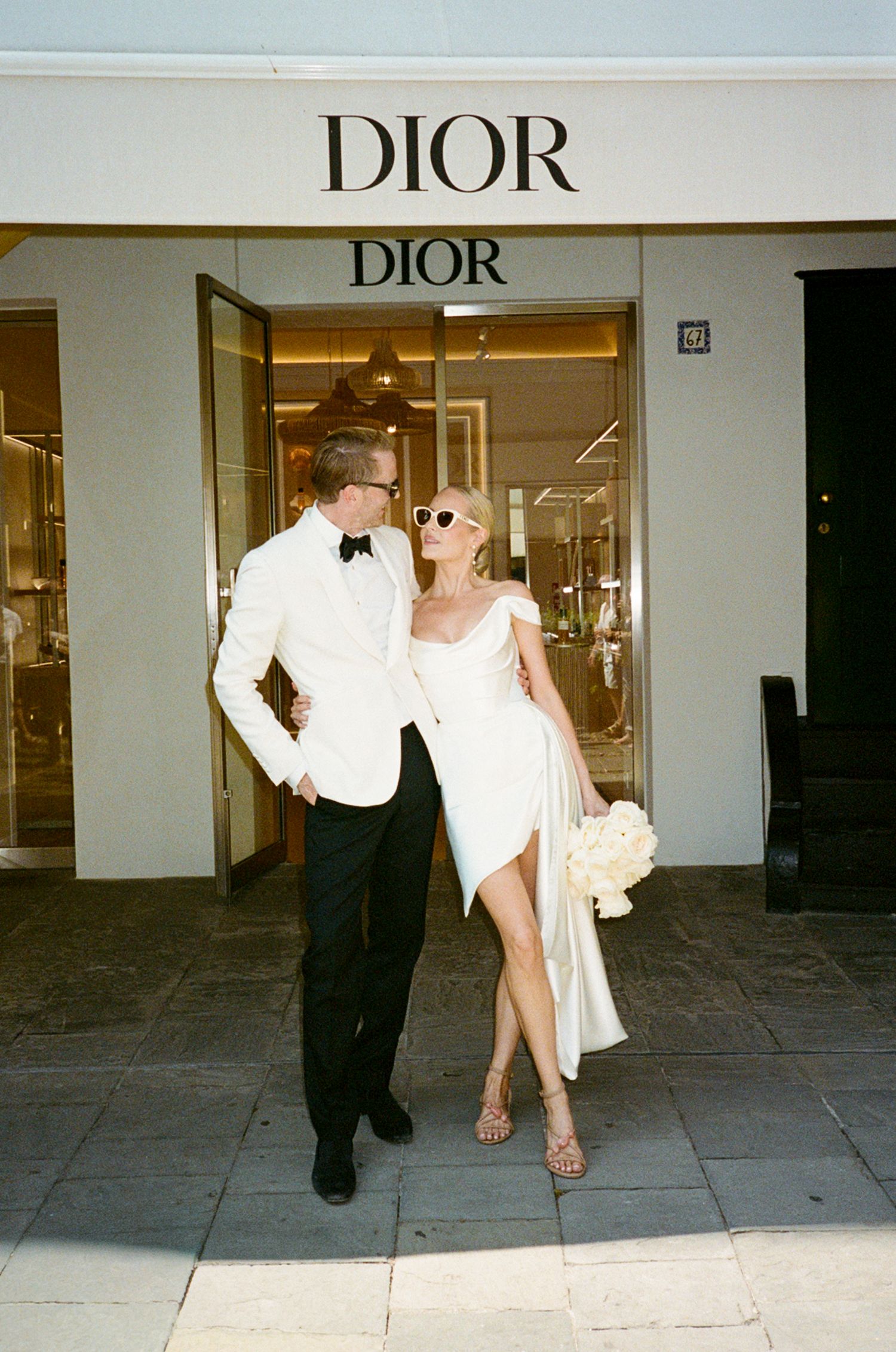 Elegant couple in white wedding attire posing outside Dior boutique on sunny day.