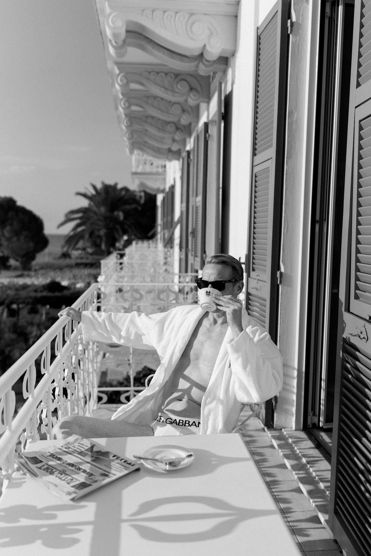 Groom in white robe drinking coffee on ornate hotel balcony in black and white photo.