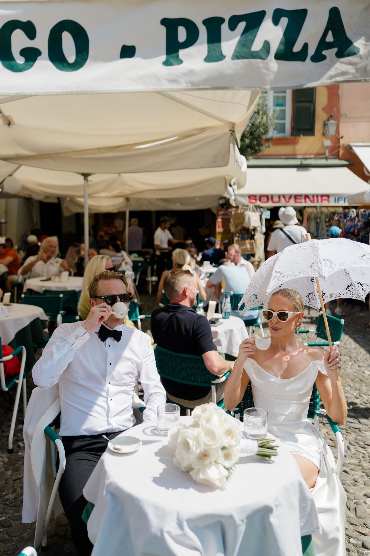 Bride and groom in sunglasses enjoying coffee at an outdoor Italian pizza cafe on their wedding day.