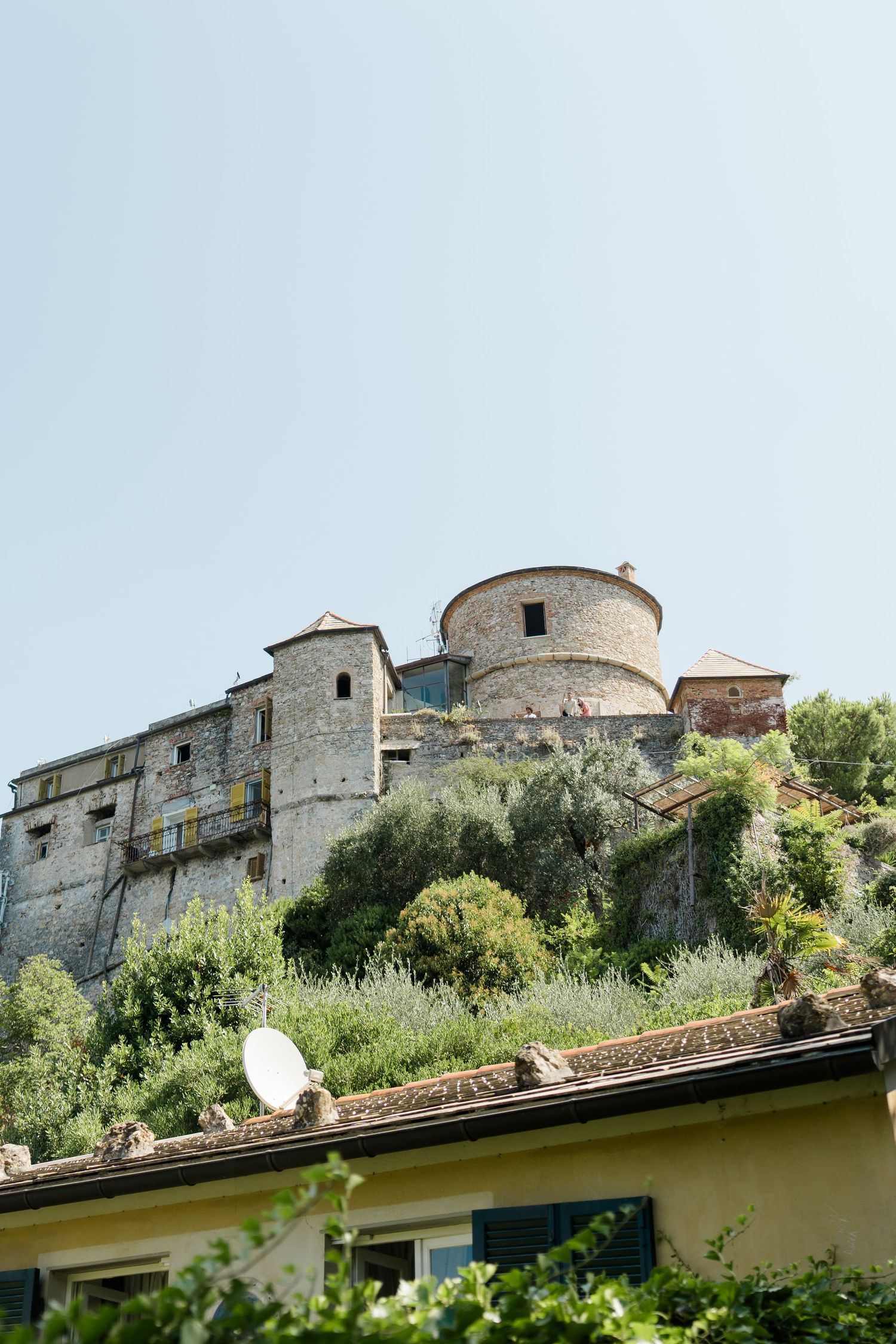 Ancient stone castle with round tower perched on hilltop surrounded by Mediterranean greenery.