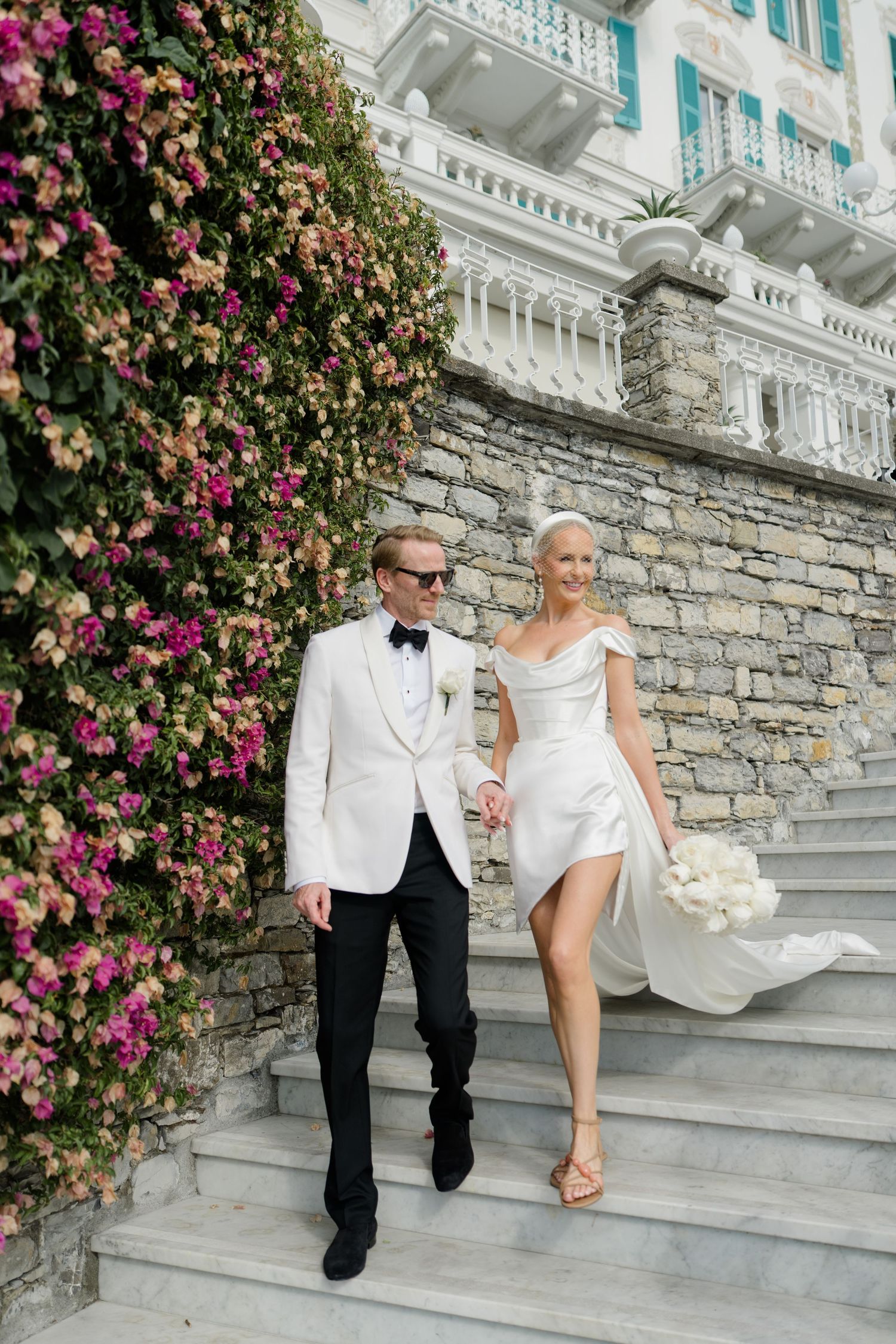 Stylish bride and groom descending stone steps beside a lush floral wall at a luxury Italian villa wedding.