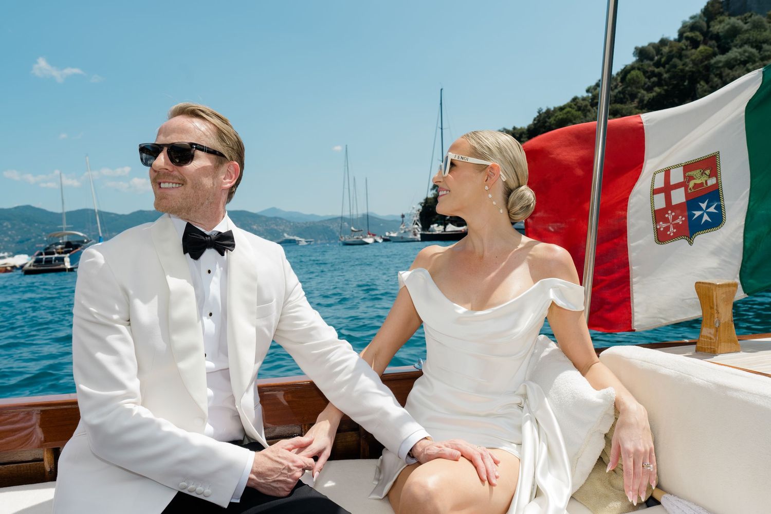 Bride and groom in elegant white attire seated on a classic wooden boat sailing on a bright blue Italian bay.