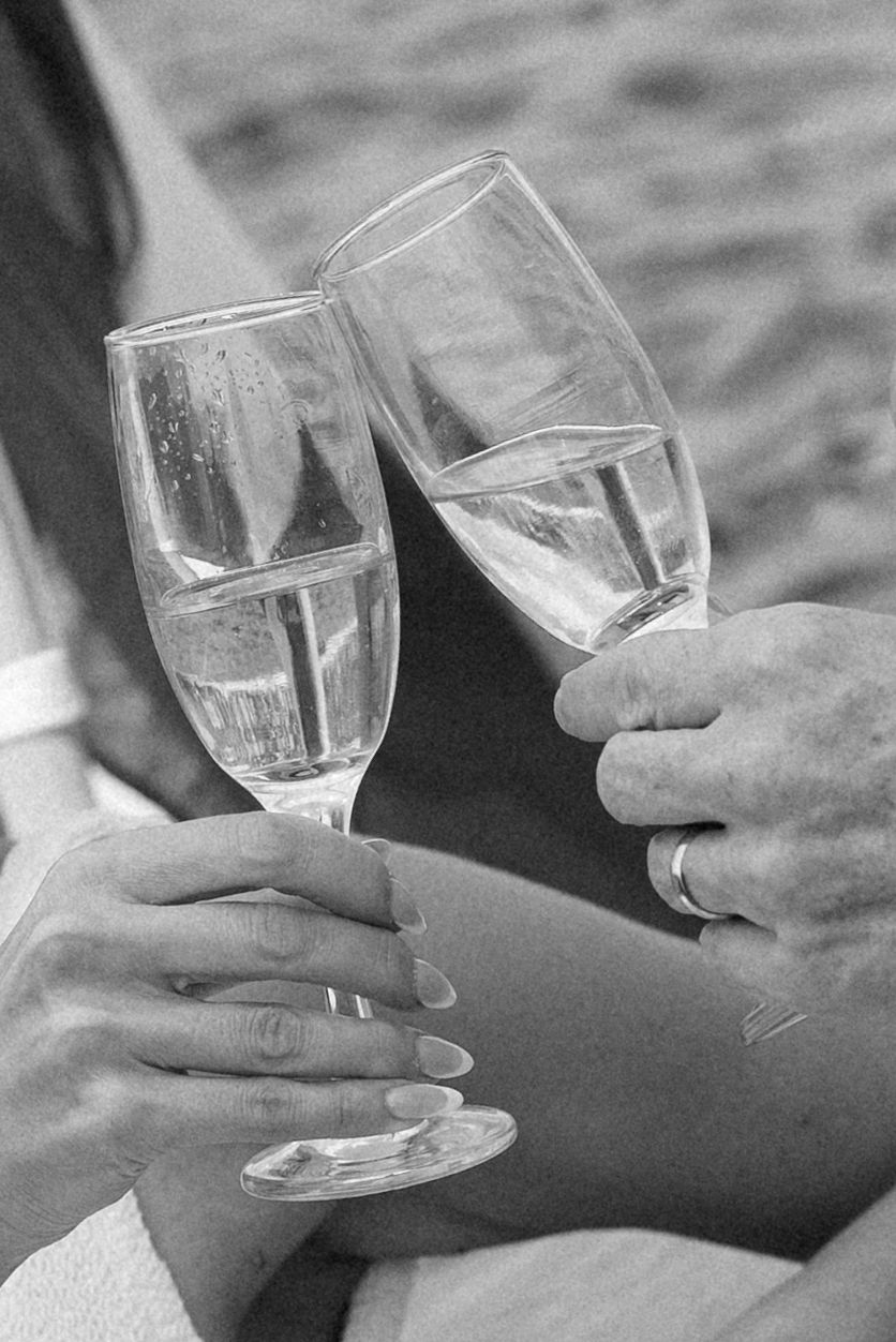 Black and white close-up of newlyweds clinking champagne flutes, wedding rings visible on their hands.