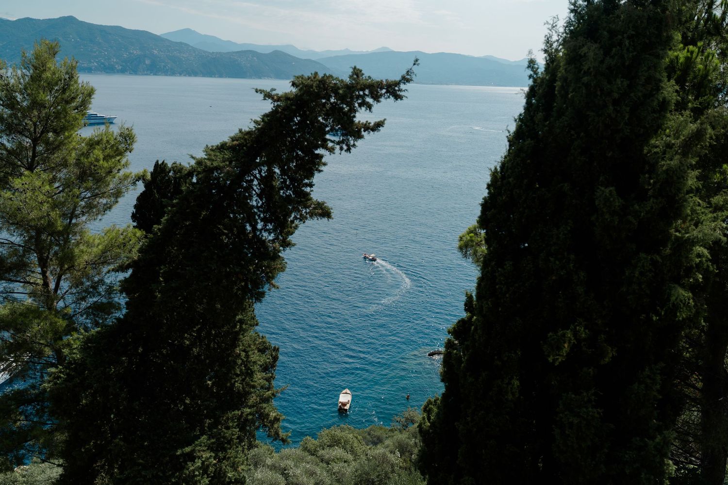 Aerial view of boats on blue Mediterranean sea framed by cypress trees with mountains in background.