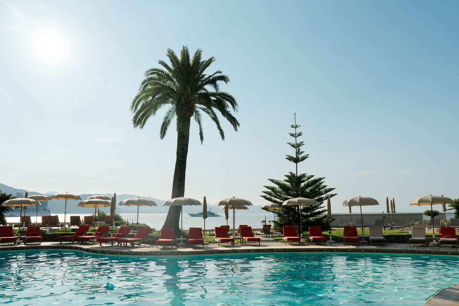 Luxury resort pool with red loungers, palm trees, and beach umbrellas under a sunny blue sky.