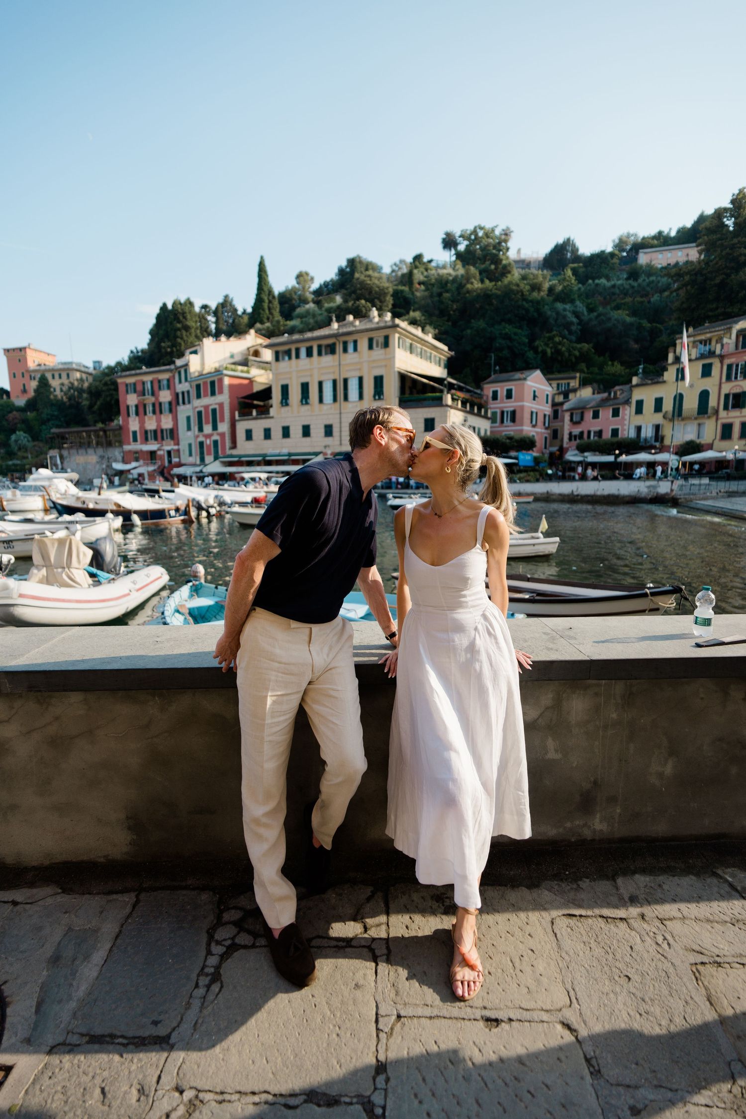 Couple sharing a romantic moment by the harbor in Portofino, Italy, with colorful buildings and boats in the background.