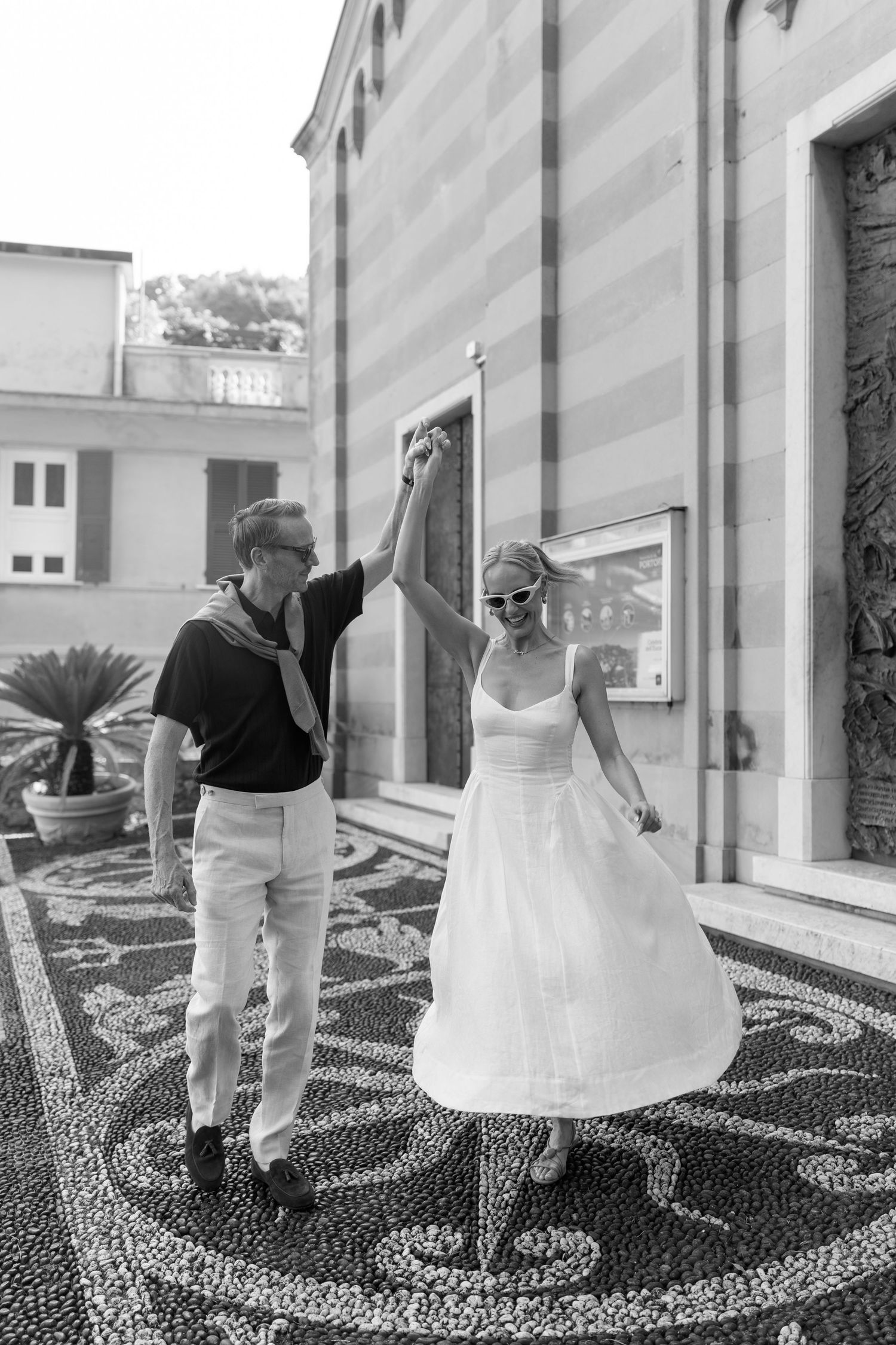 Black and white photo of couple dancing on mosaic plaza outside Italian church, woman in white dress twirling.