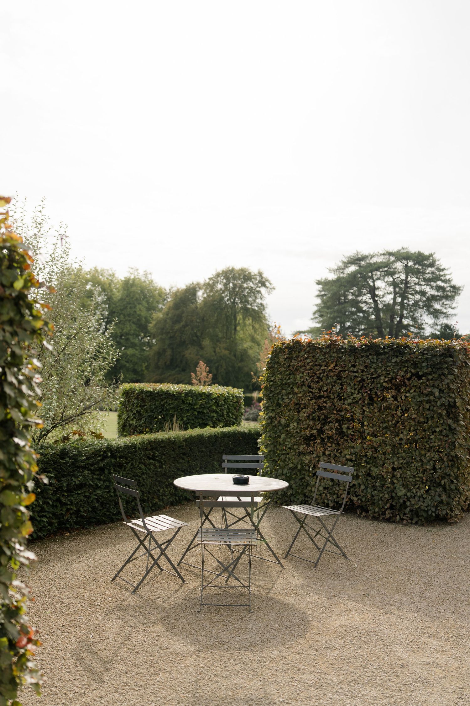 A serene garden patio with metal bistro table and chairs surrounded by neatly trimmed hedges and trees.