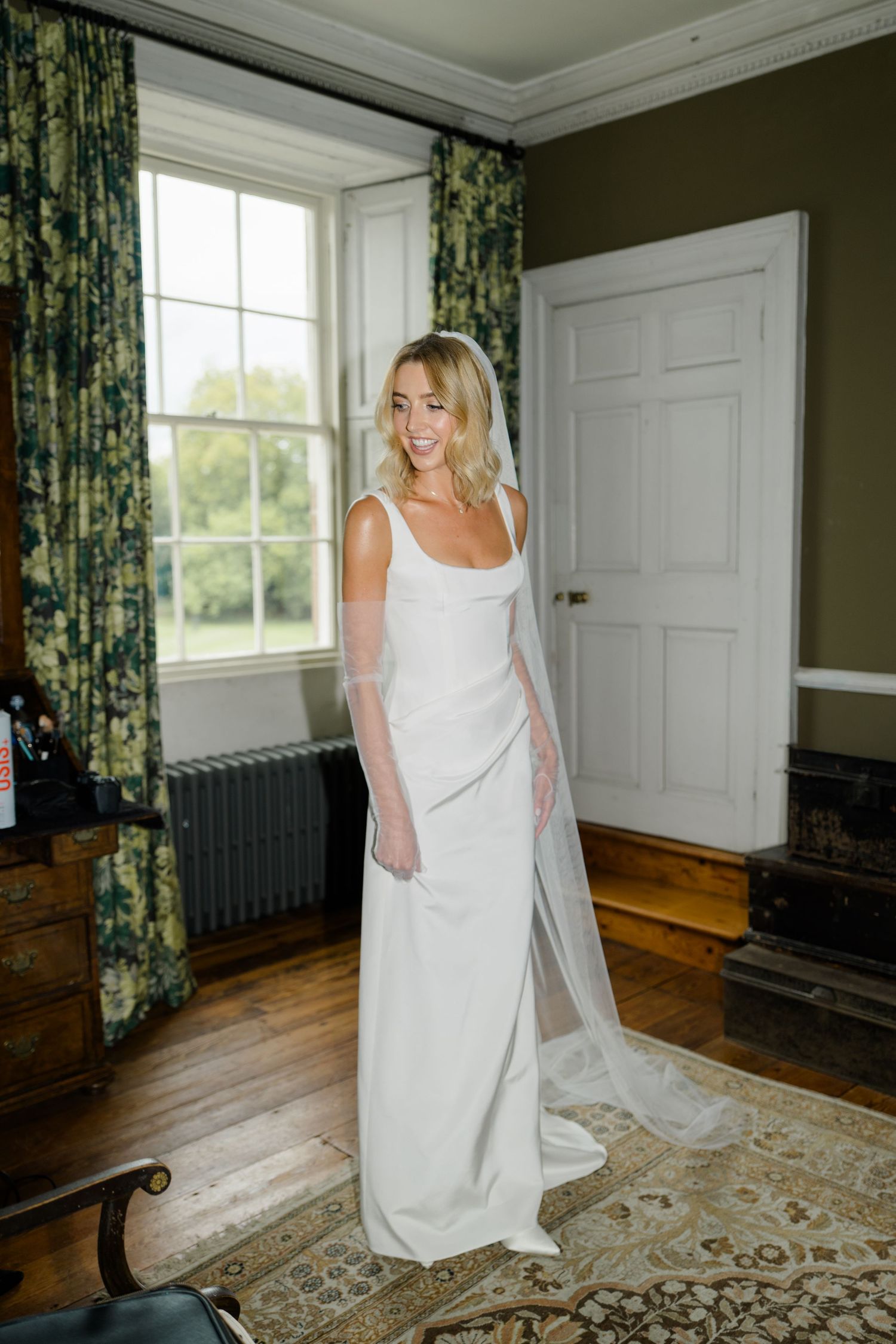 Bride in simple white wedding dress with long veil standing in classic room.