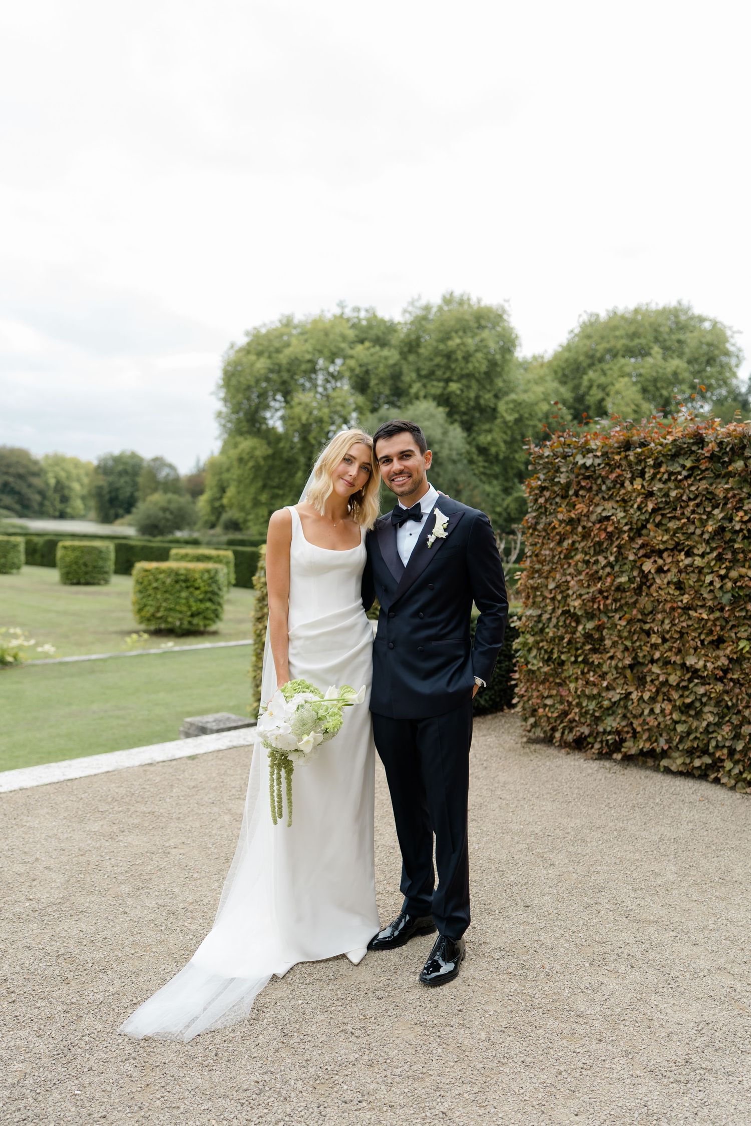 Smiling bride and groom pose together in formal wedding attire in manicured garden grounds.