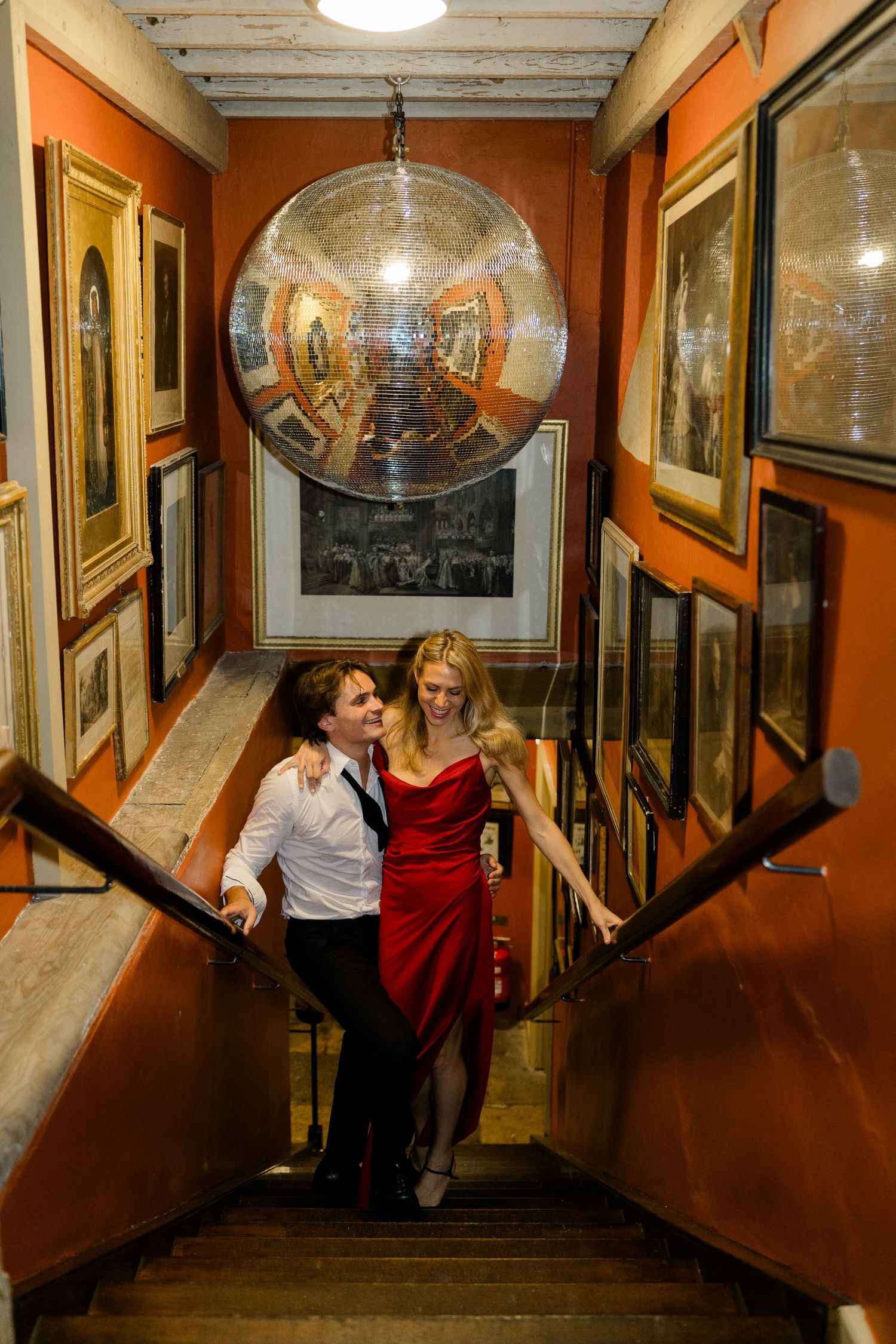 Couple in formal attire walking up stairs lined with artwork beneath a disco ball in elegant venue.