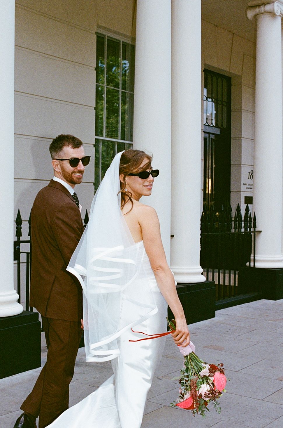 Bride and groom in sunglasses walking outside a white building, bride holding a pink bouquet.