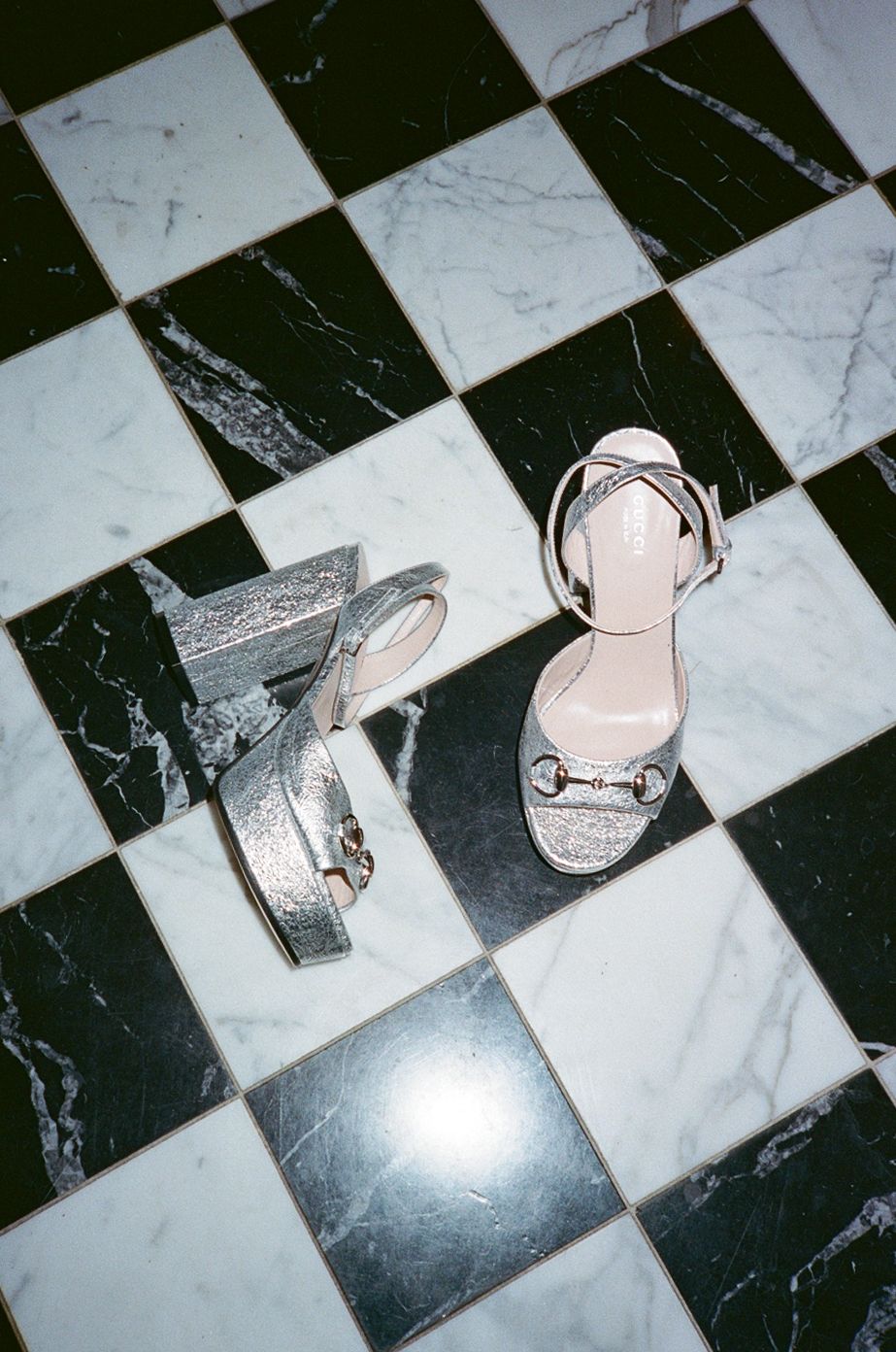 Silver glitter platform wedge and strappy heeled sandals displayed on black and white marble checkered floor.