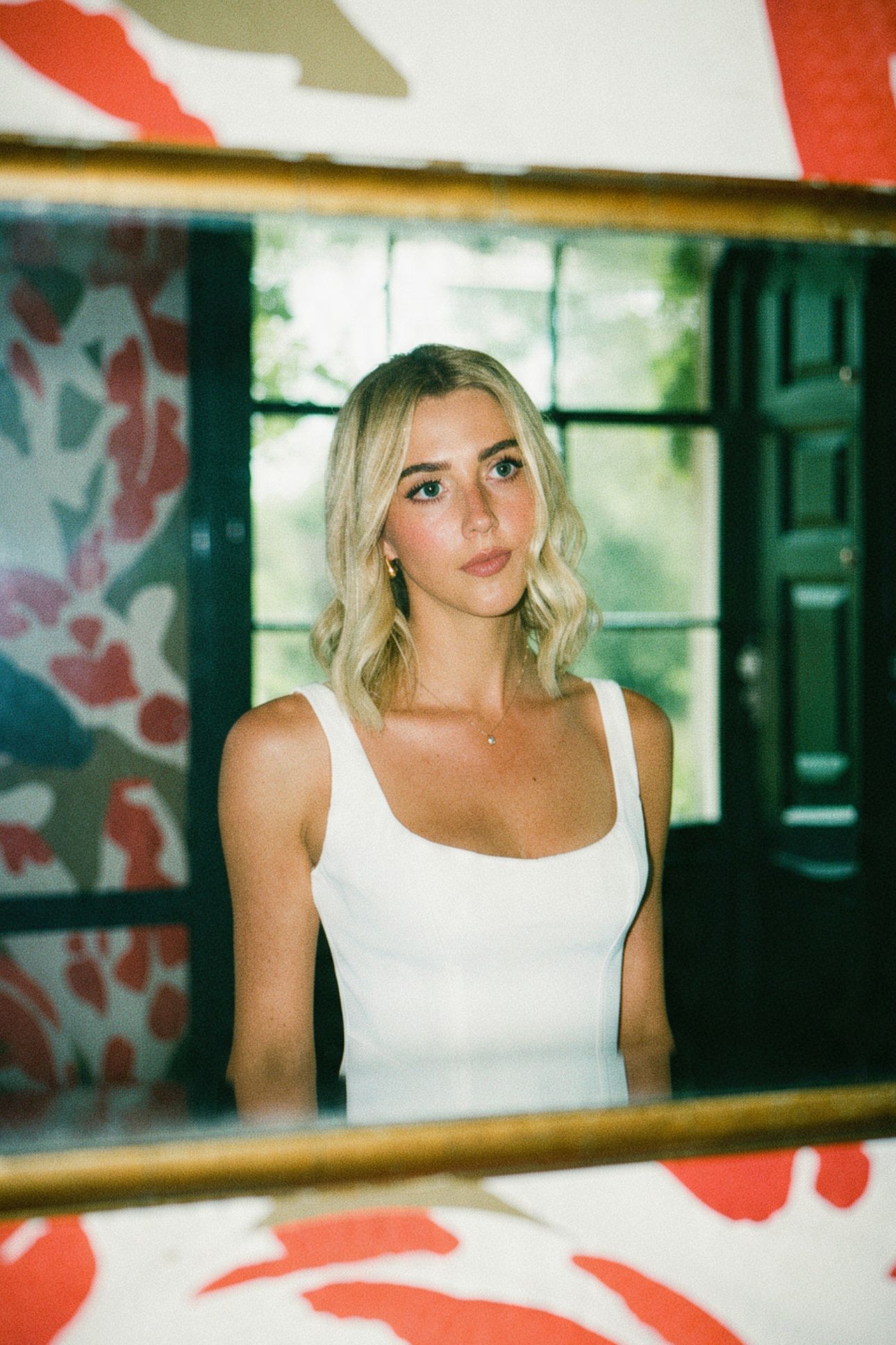 Young blonde woman in white tank top reflected in gold-framed mirror with floral wallpaper background.