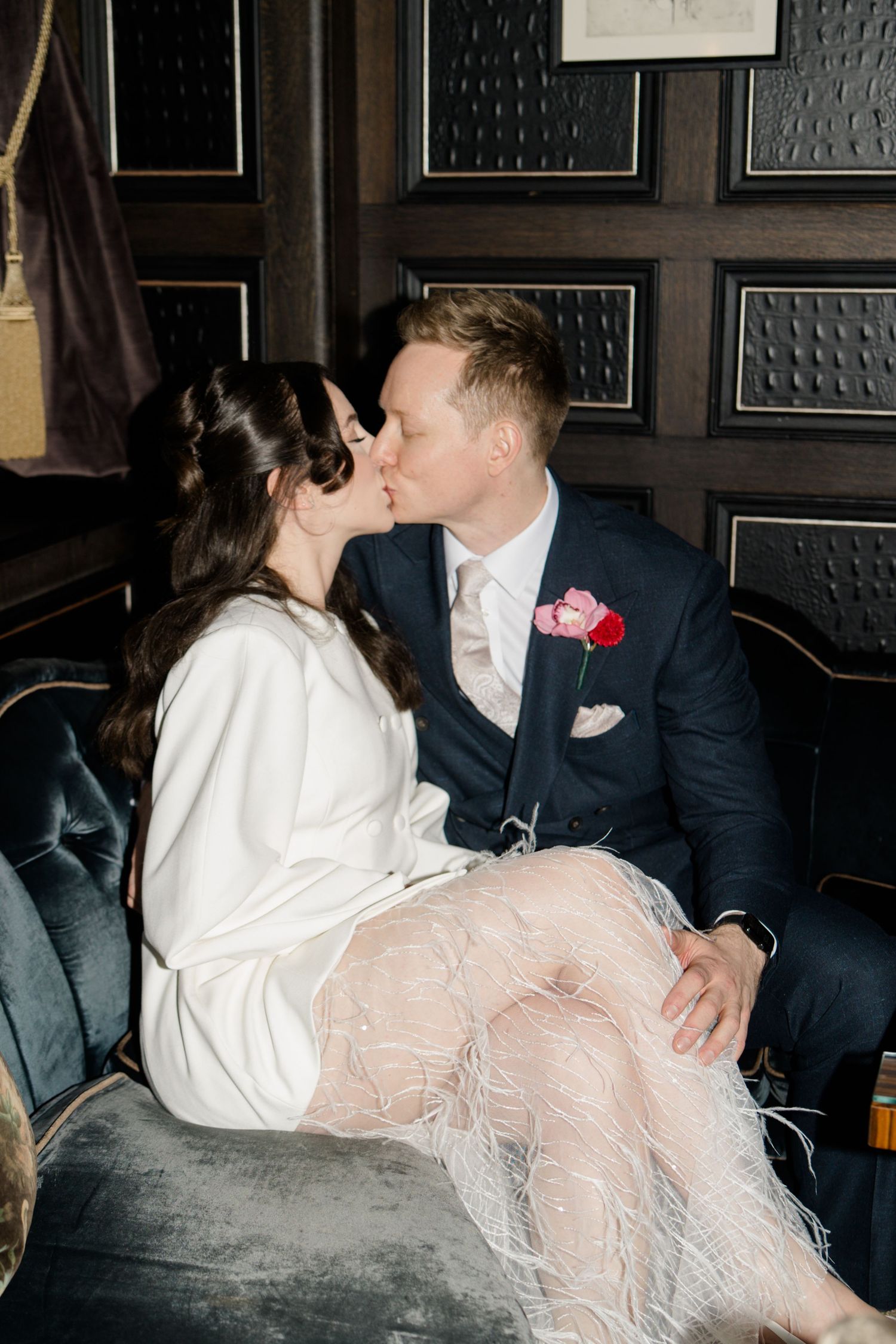 Bride in white feathered skirt and groom in navy suit share a kiss on a velvet sofa.