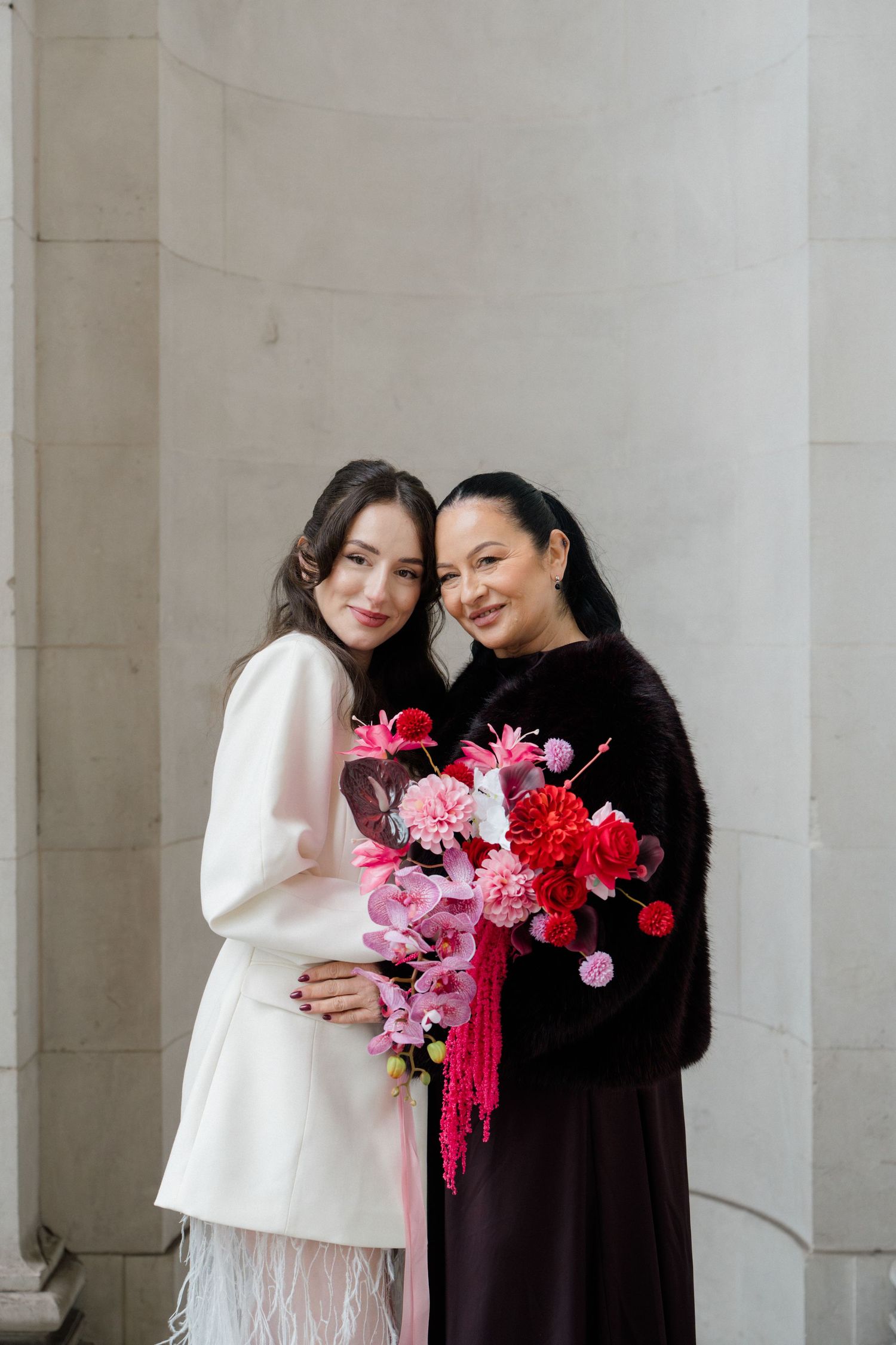 Bride in white blazer and mother in black fur coat holding vibrant pink and red floral bouquets together.