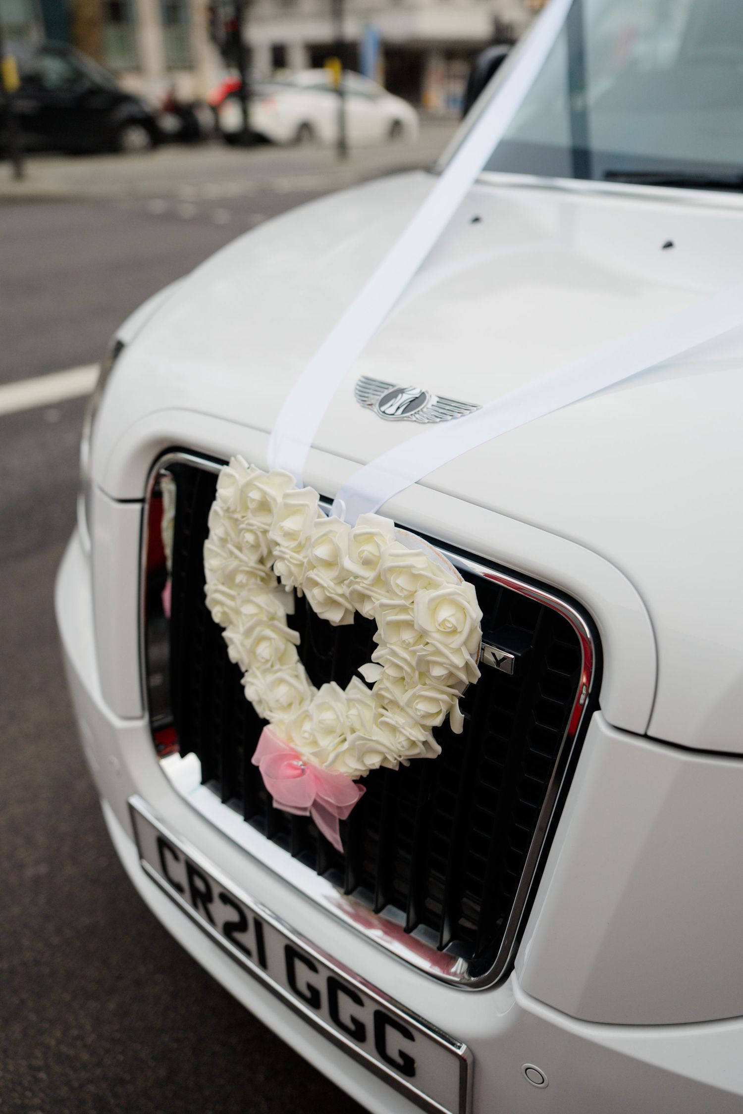White wedding car decorated with heart-shaped white rose wreath and ribbon on hood.